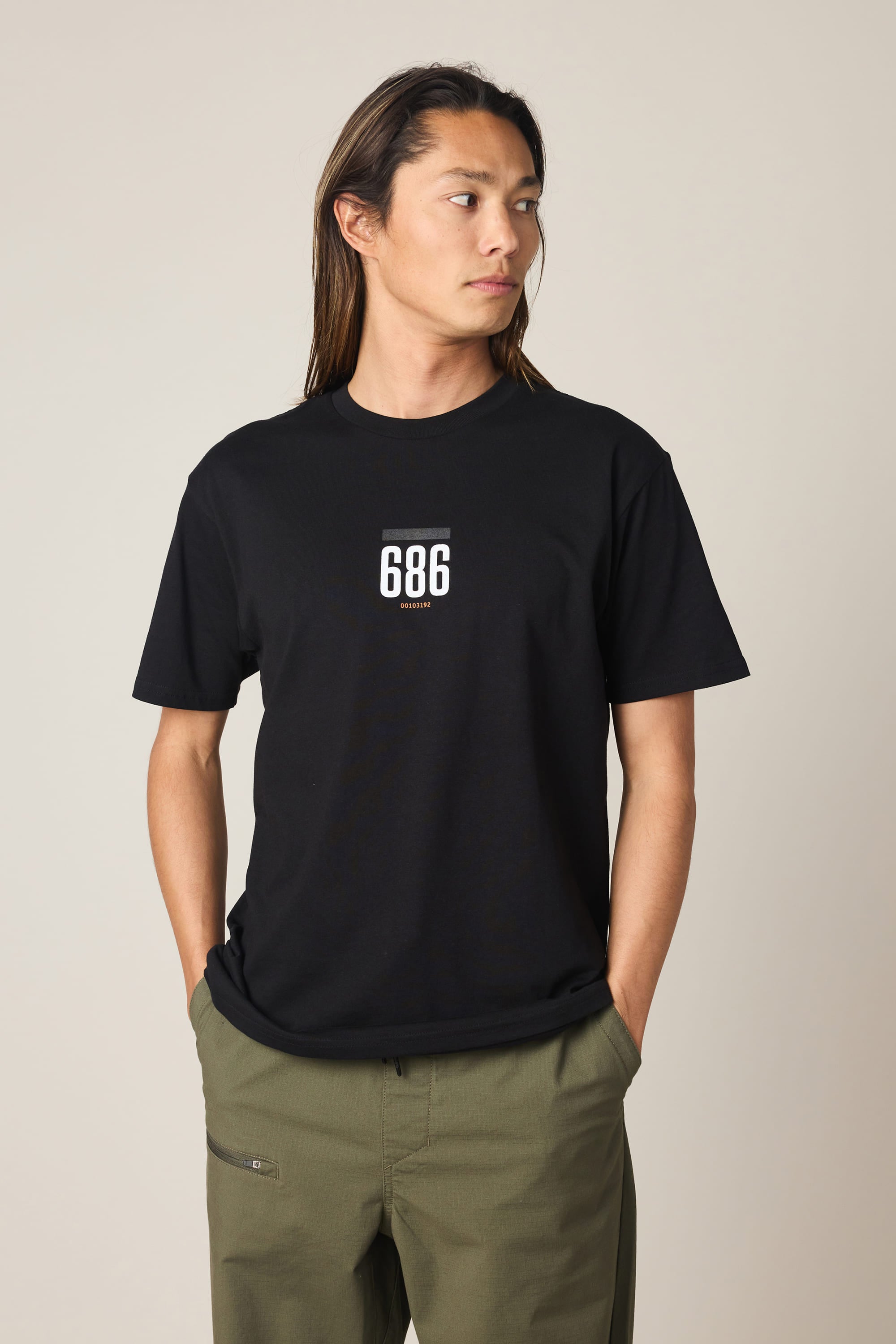 A person with long hair stands against a plain background, wearing the 686 Lifts Authentic SS Tee in black by 686, featuring “686” in white on the chest, paired with olive green pants and hands in pockets, looking to the side.
