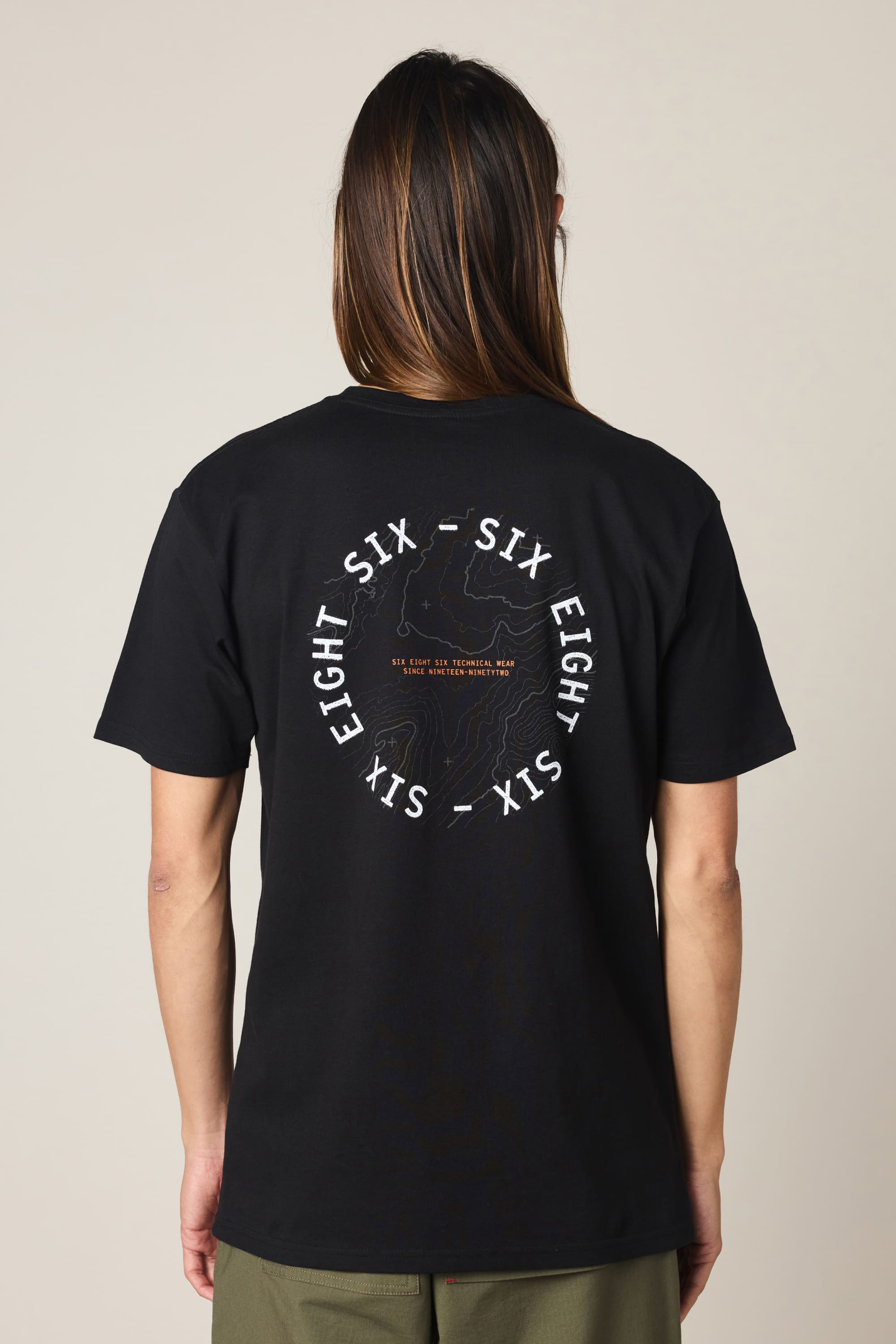 A person with long brown hair wears the 686 Lifts Authentic SS Tee by 686, a men's black cotton t-shirt with "SIX EIGHT SIX" in white circular text and a line art graphic on the back, set against a plain neutral background.