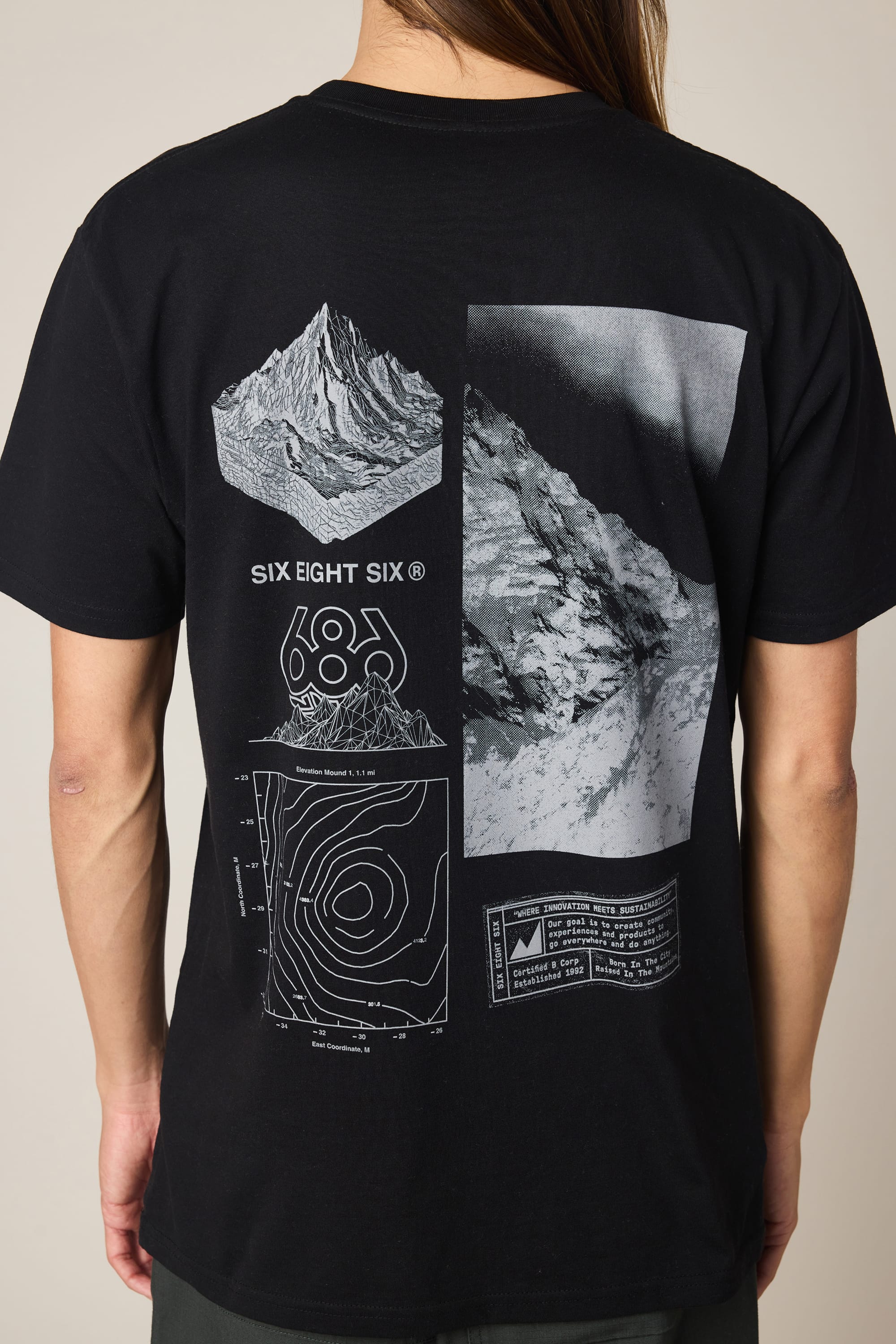 A person with long hair is seen from behind wearing the 686 Cross-Cut Authentic S/S Tee by 686—a black 100% cotton T-shirt featuring white graphic designs of a mountain, topographic map, and "SIX EIGHT SIX" text on the back.