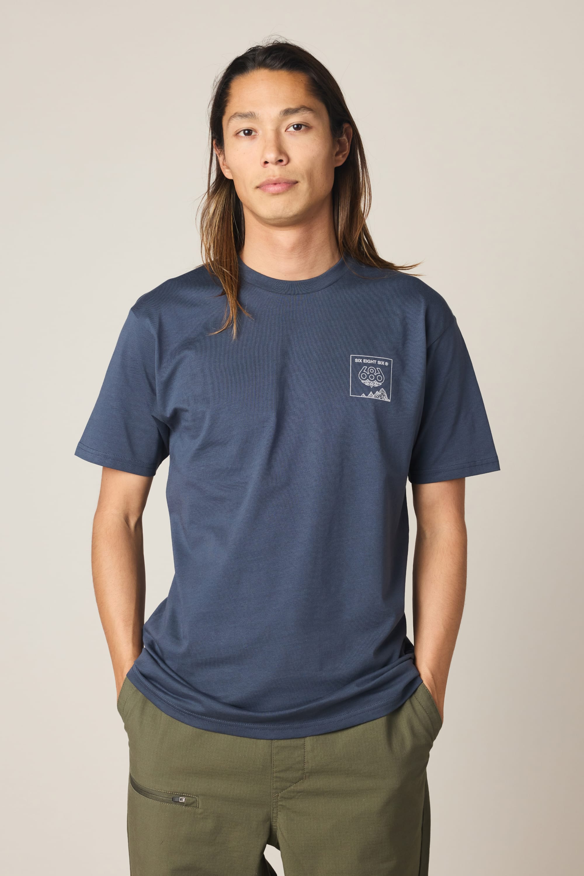 A person with long hair wears the 686 Cross-Cut Authentic S/S Tee by 686 in navy blue with a small white chest graphic and olive green pants, standing against a plain light background.