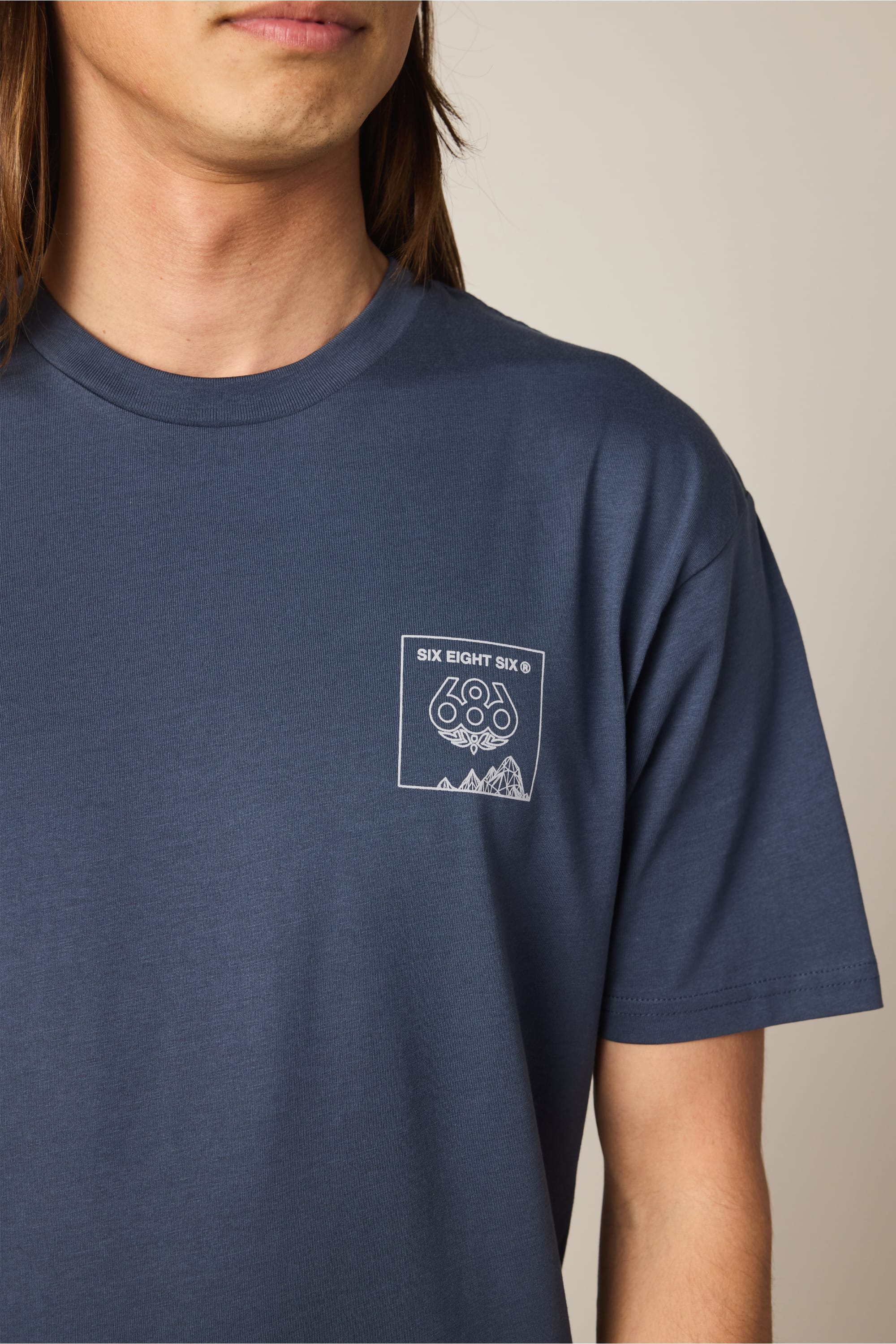 A person wears the 686 Cross-Cut Authentic S/S Tee, a blue men's cotton t-shirt by 686 with a small chest logo displaying "SIX EIGHT SIX," geometric shapes, and mountain outlines. Only their lower face, neck, and upper torso are visible.