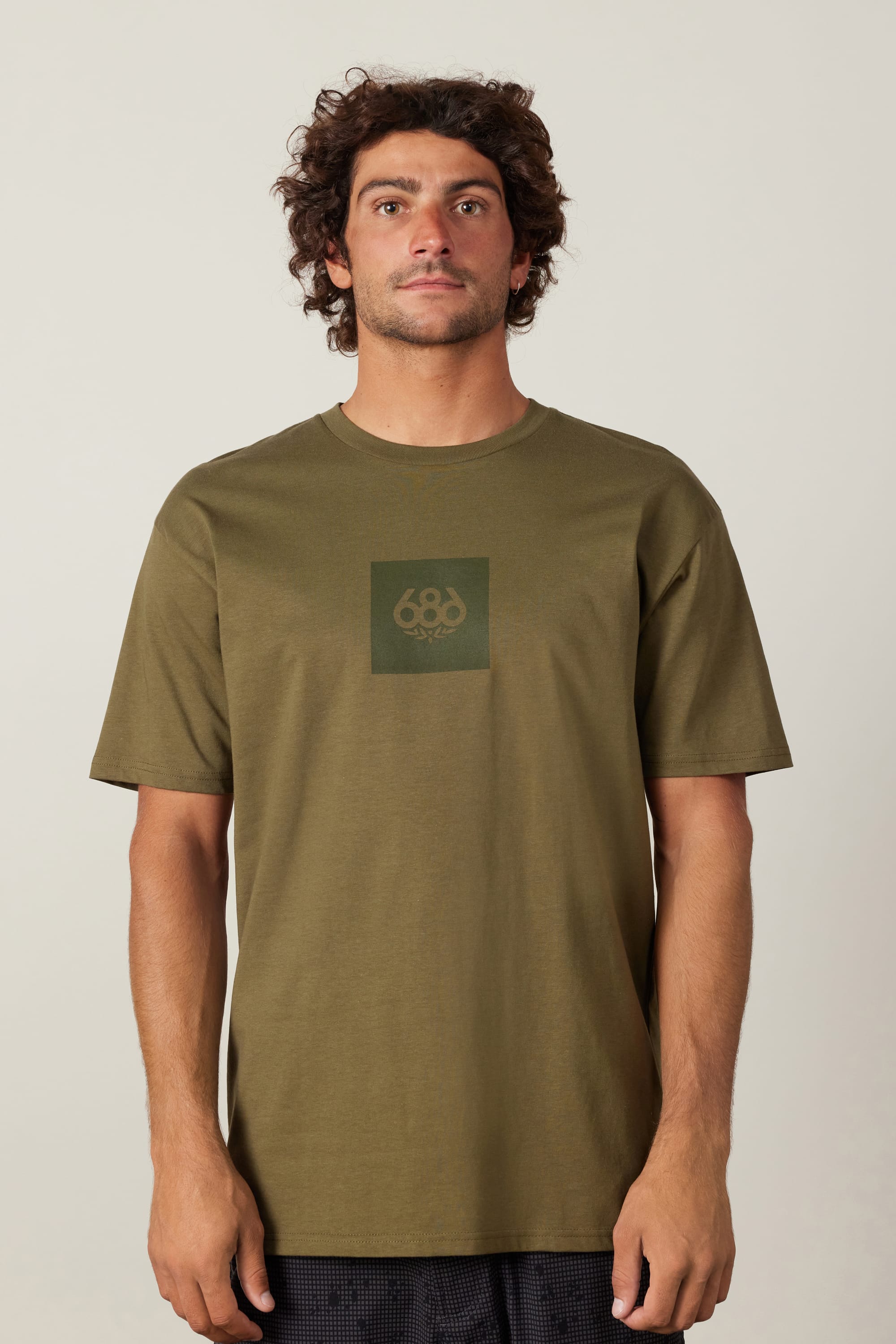 A man with curly brown hair wears the 686 Knockout Authentic S/S Tee, an olive green 100% cotton men's t-shirt with a subtle 686 logo on the chest, standing facing the camera against a plain light background.