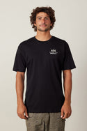 A man with curly hair wears the 686 Path Authentic S/S Tee, a black 100% cotton men’s t-shirt with “686” and white text on the left chest, paired with patterned khaki shorts. He stands facing forward against a plain light background.
