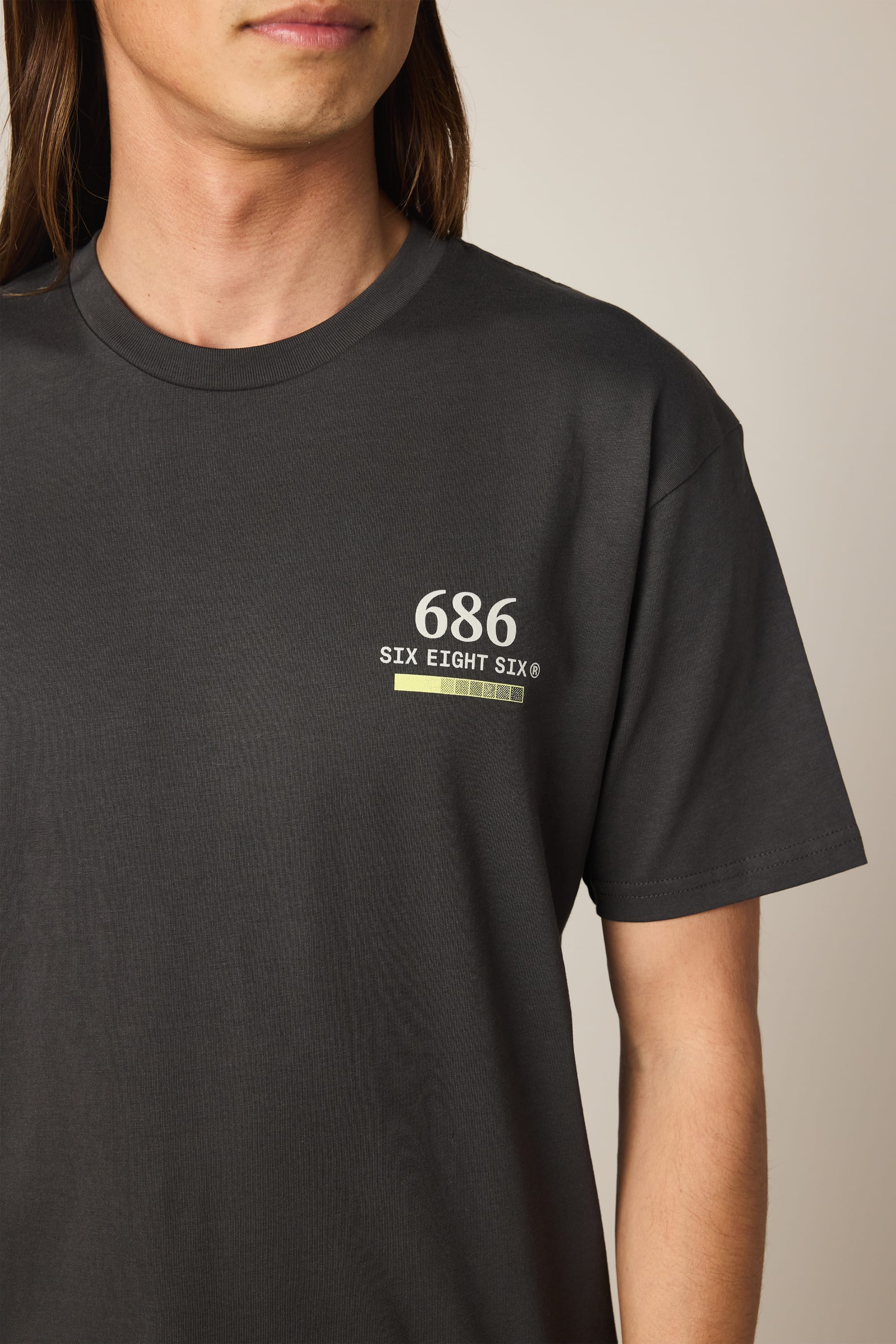 A person with long brown hair is wearing the 686 Path Authentic S/S Tee by 686, a black men’s t-shirt featuring “686 SIX EIGHT SIX” printed in white on the upper left chest. The shirt is made from 100% cotton, and only the upper torso and part of the face are visible.