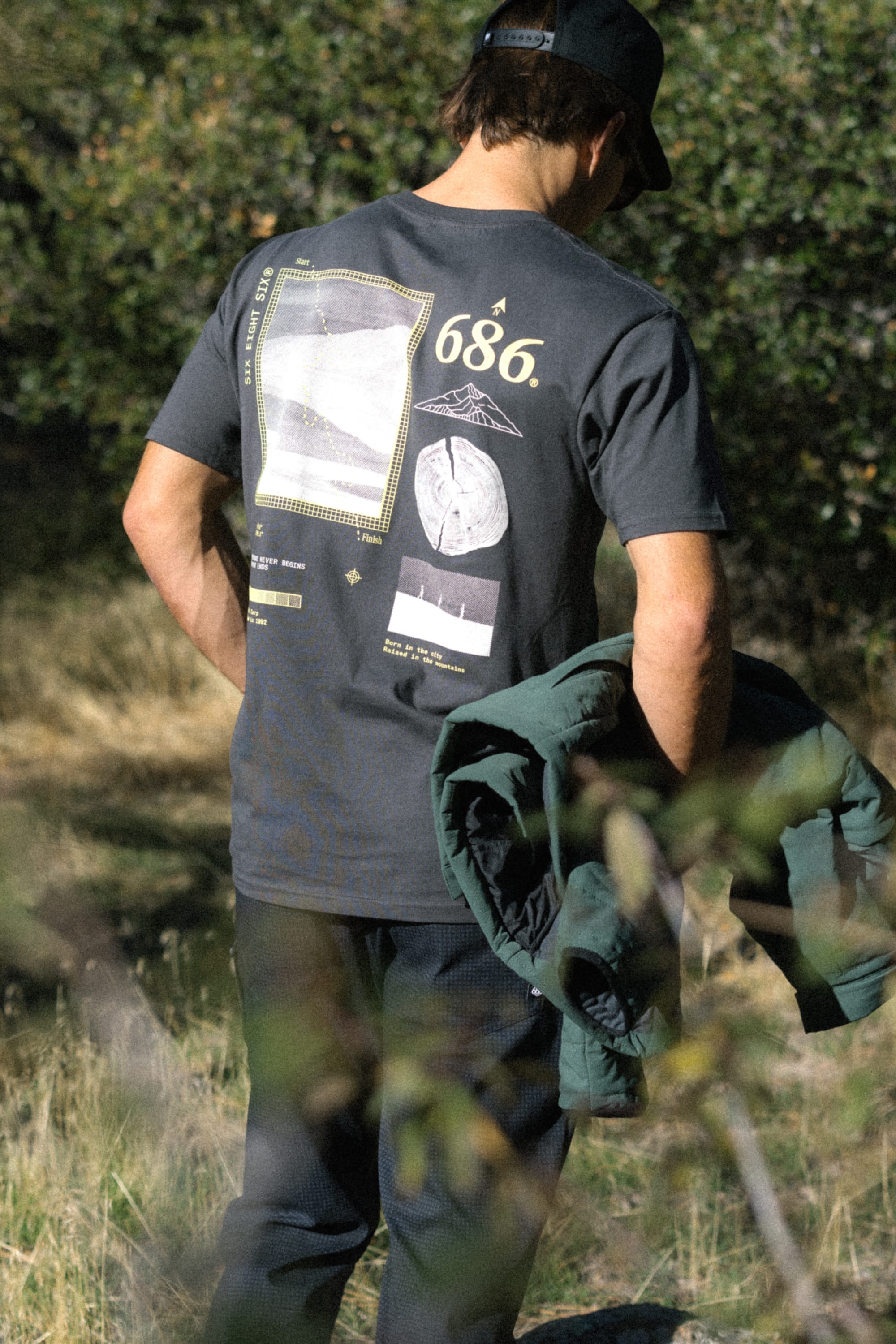 A person stands outdoors with their back to the camera, holding a green jacket. They are wearing the 686 Path Authentic S/S Tee by 686—a men's graphic t-shirt made of 100% cotton—paired with dark pants and a black cap. Green foliage and grass fill the background.