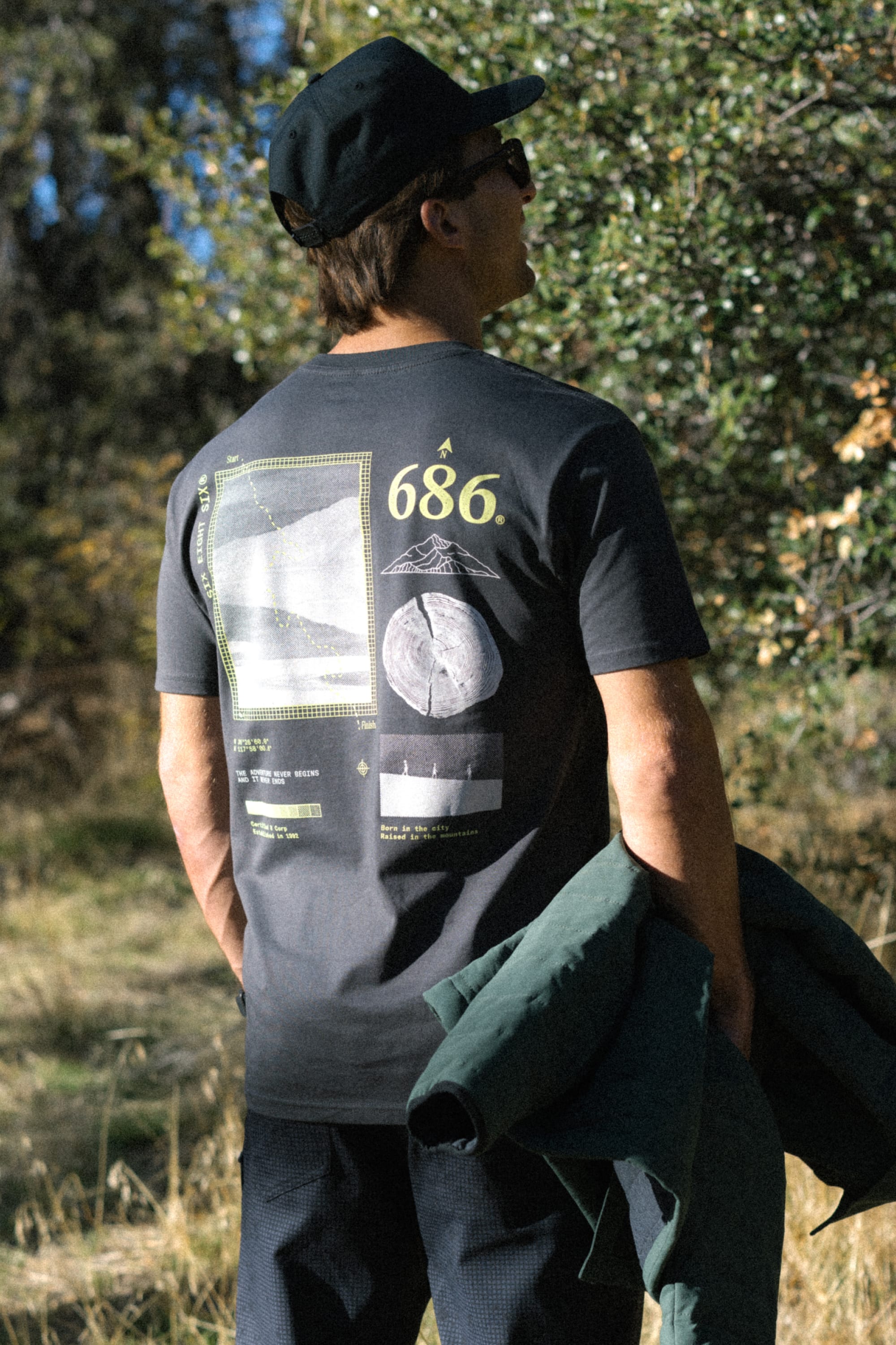 A person wearing a black cap and the 686 Path Authentic S/S Tee by 686 stands outdoors, facing away and holding a green jacket, as sunlight filters through the trees in the background.