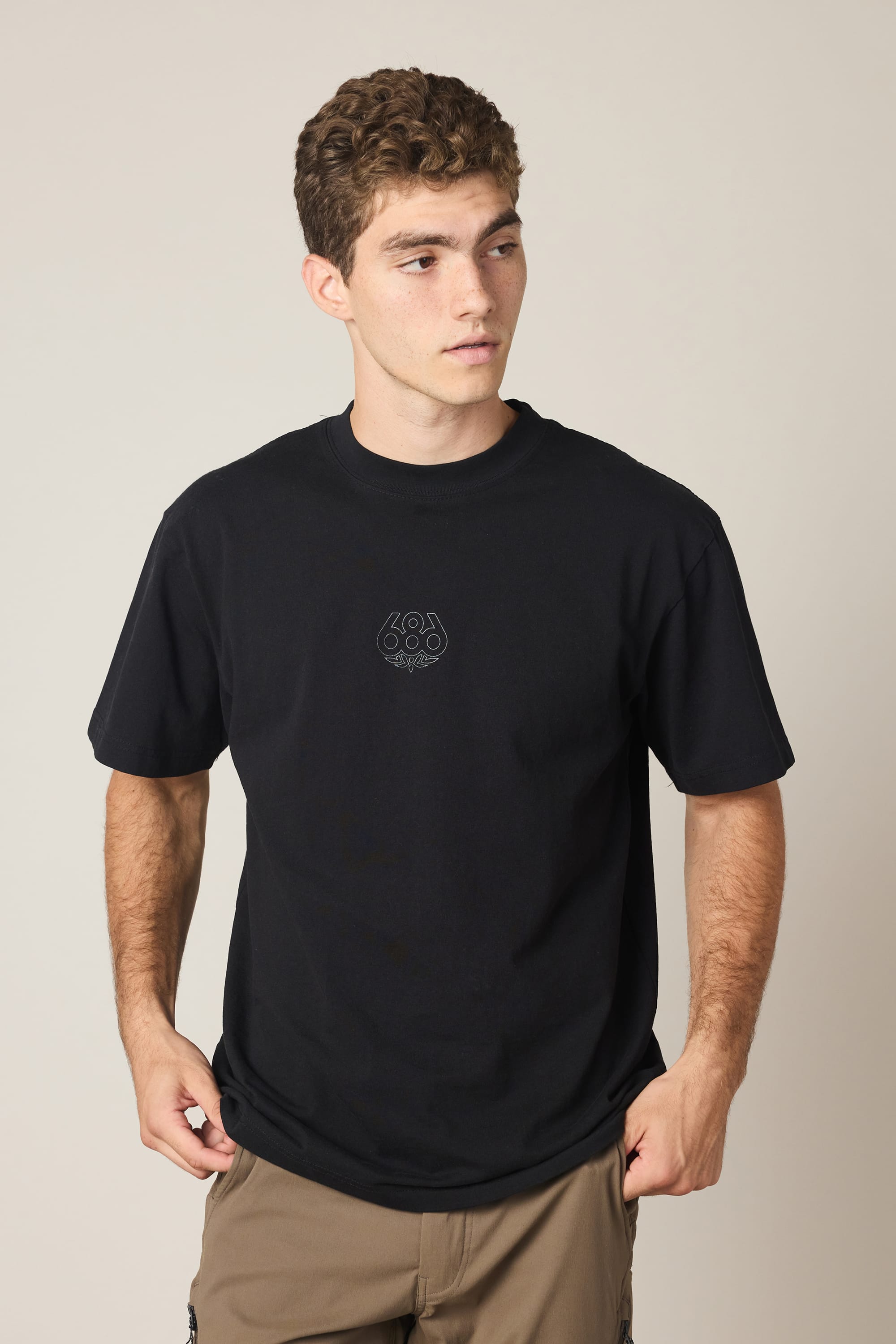 A young man with short curly hair is wearing the 686 High Def Relaxed S/S Tee by 686—a black men’s t-shirt made from 100% cotton with a small logo—paired with tan pants. He stands against a plain light background, looking slightly to his left.