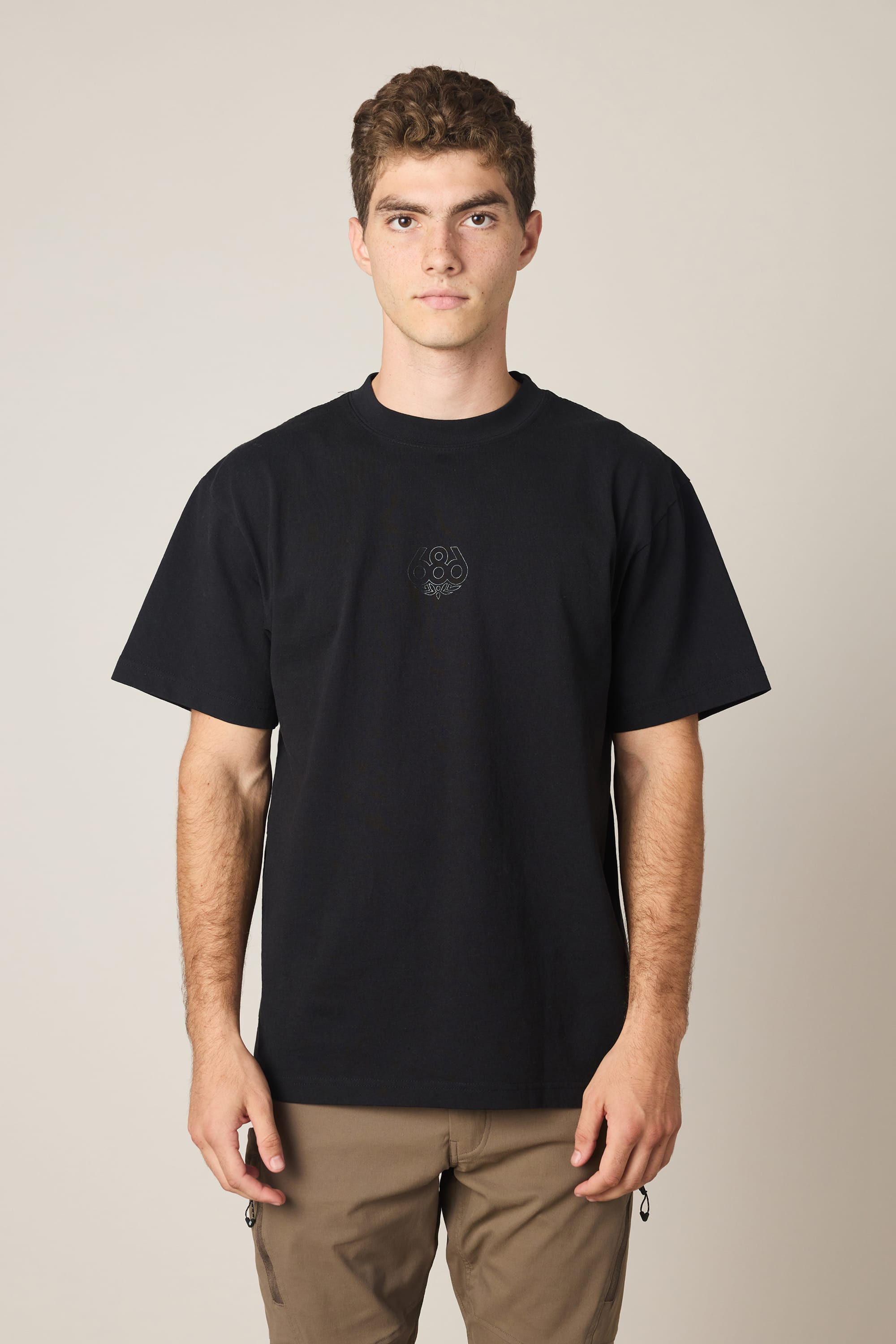 A young man with short curly hair wears the 686 High Def Relaxed S/S Tee by 686, a plain black men's t-shirt made from 100% cotton, paired with tan pants. He stands against a neutral background, looking directly at the camera with a neutral expression.