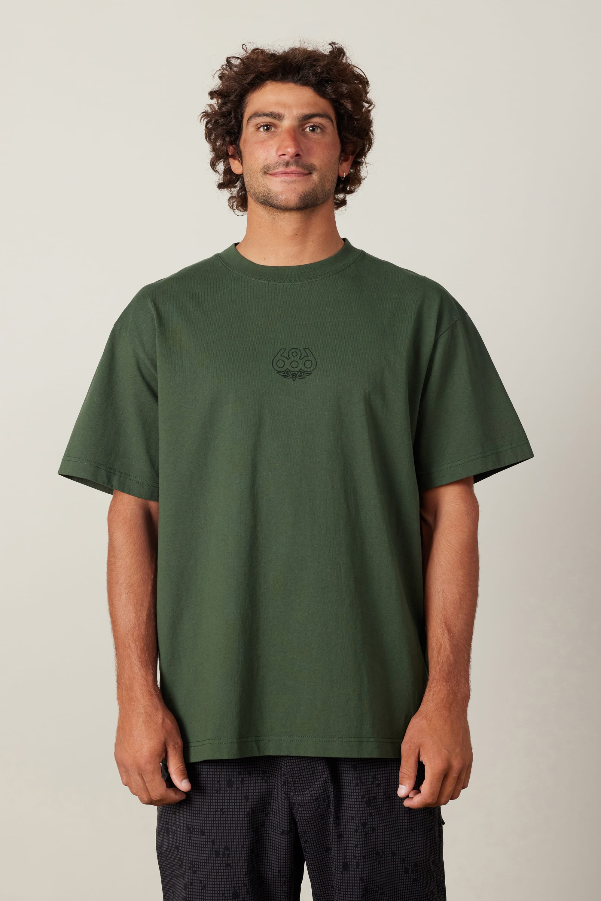 A man with curly hair wears the 686 High Def Relaxed S/S Tee by 686, a loose-fitting dark green men's cotton t-shirt with a small logo on the chest, paired with black textured shorts, standing against a plain light background.