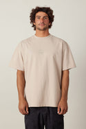 A man with curly brown hair wears the 686 High Def Relaxed S/S Tee by 686 in beige, paired with dark patterned shorts, standing facing forward against a plain light background.
