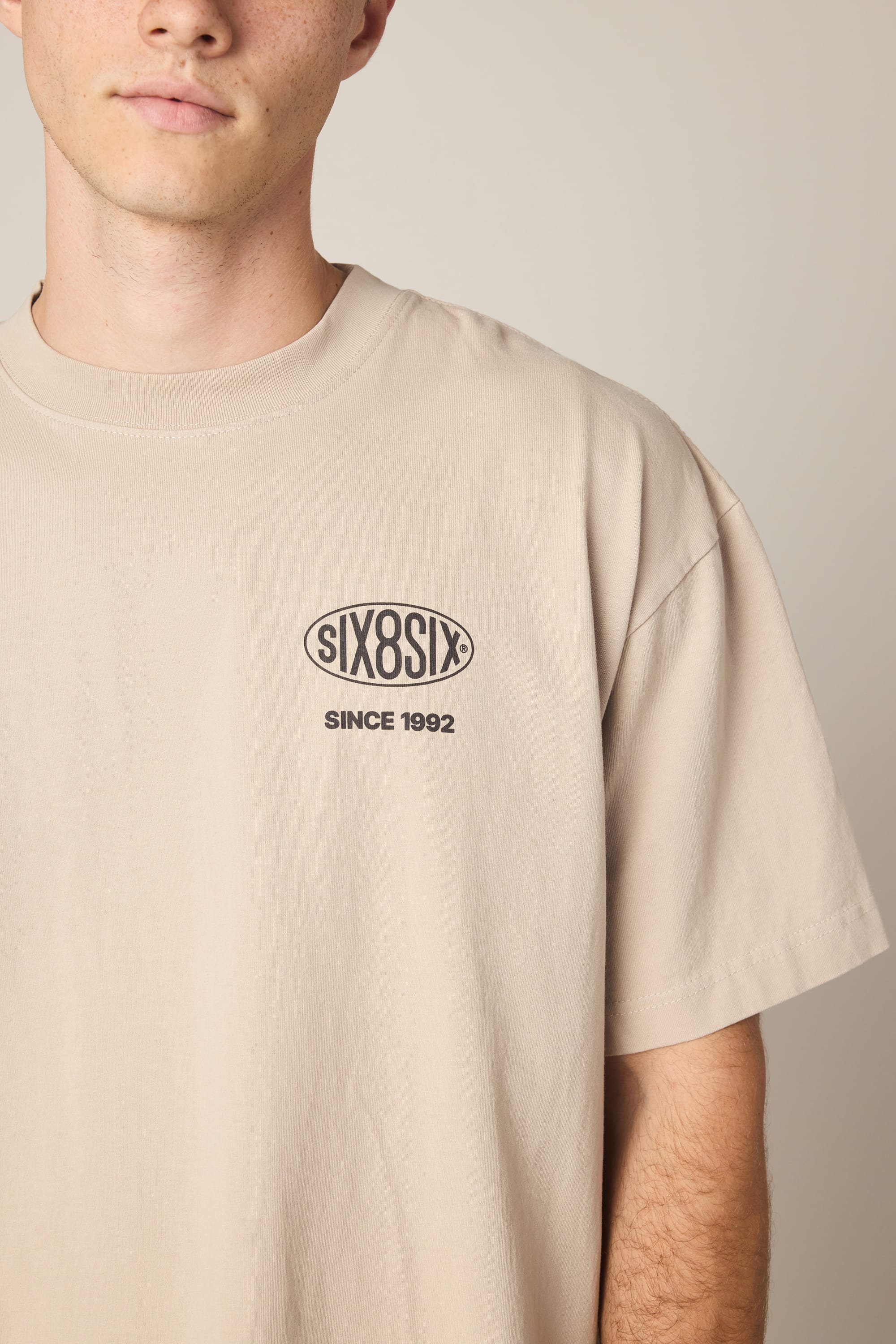 A person wearing the 686 92 Relaxed S/S Tee—a beige men's T-shirt from 686 featuring the SIX8SIX logo and "SINCE 1992" text on the left chest. Made from 100% cotton, only the lower half of their face and upper body are visible.
