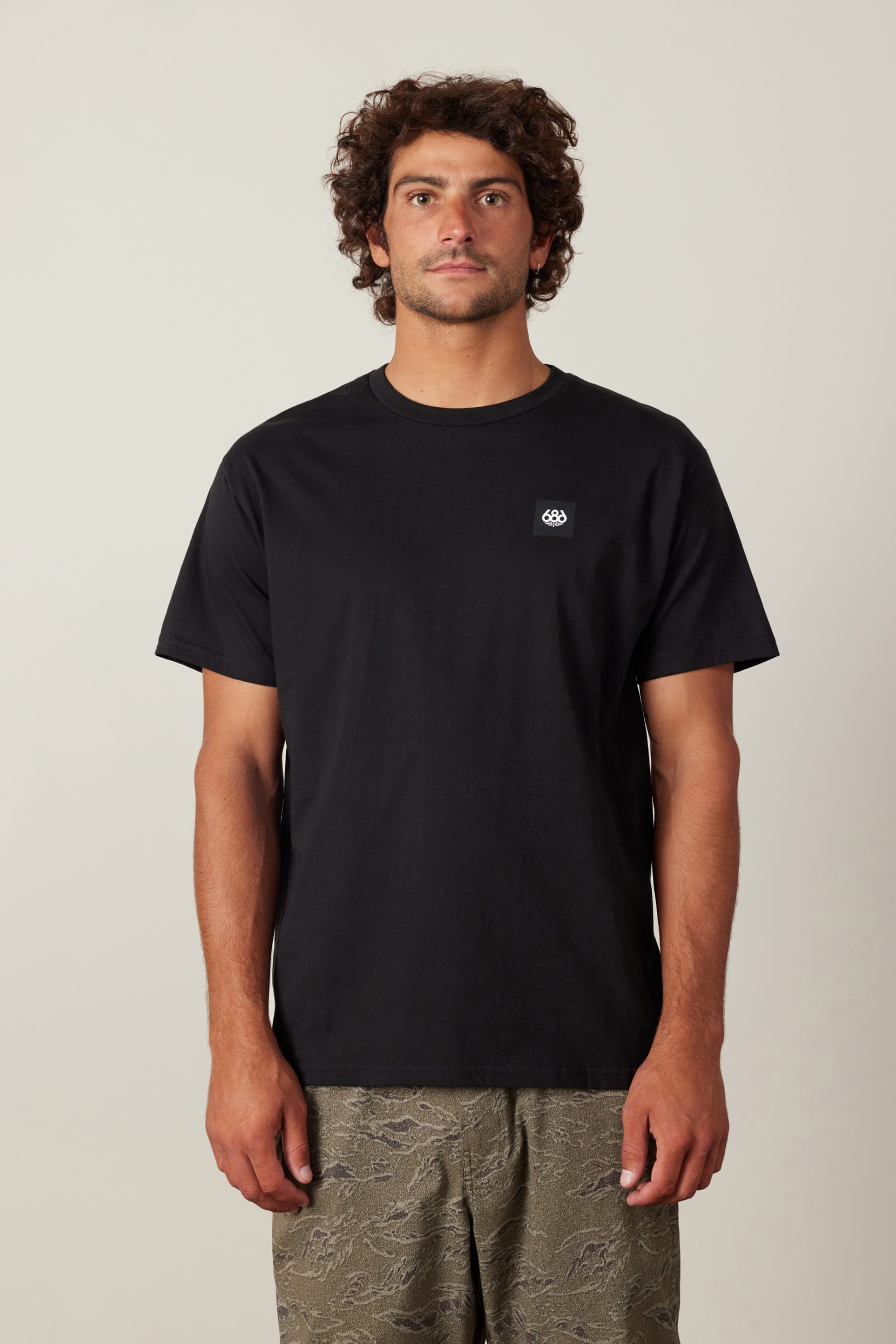 A man with curly brown hair wears the 686 Logo 3-Pack S/S Tee by 686, a plain black 100% cotton men's t-shirt with a small white logo on the left chest, paired with patterned green shorts against a light background.