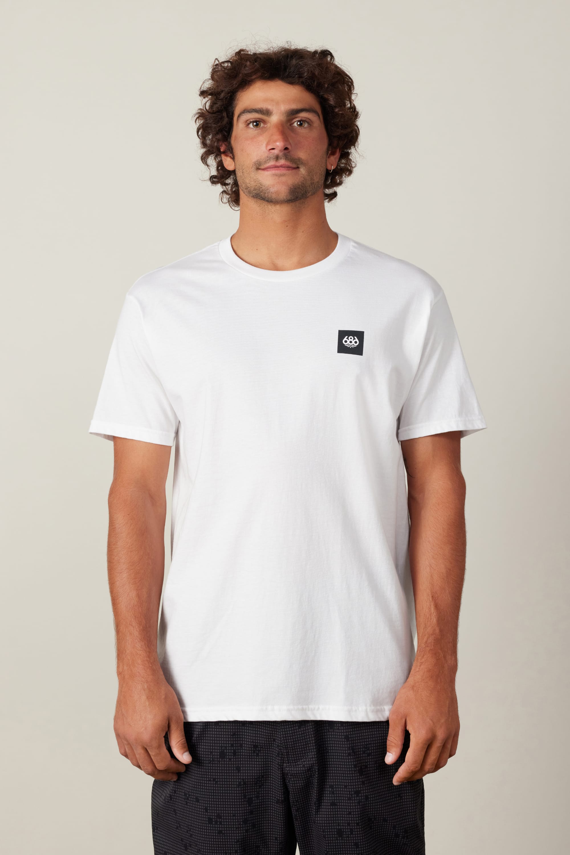 A man with curly brown hair wears a plain white 686 Logo 3-Pack S/S Tee by 686 featuring a small black logo on the left chest and black patterned shorts, standing against a light background.