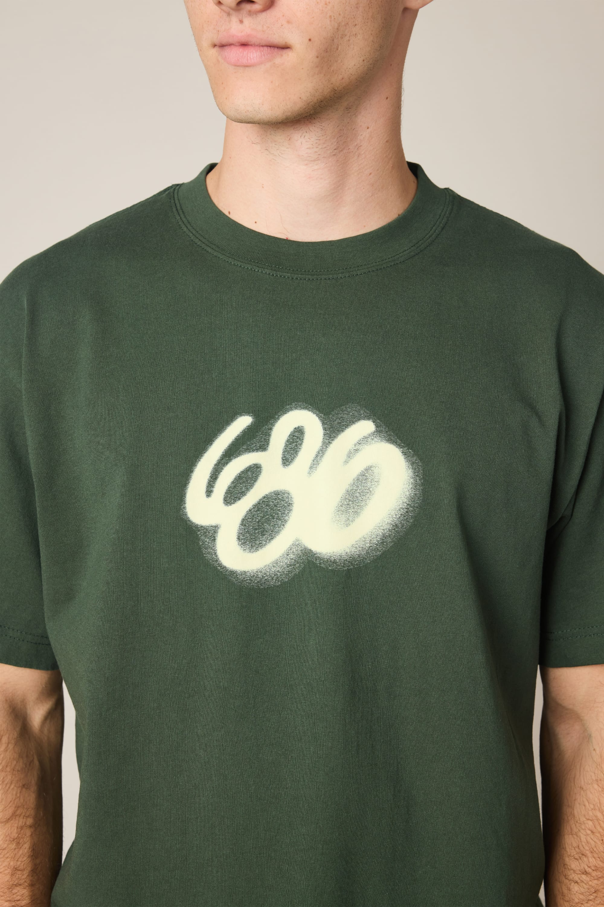A person wearing the 686 Spray Relaxed SS Tee—a dark green men’s t-shirt featuring a white, spray-painted 686 logo. Only the lower face and upper torso are shown, emphasizing its 100% cotton fabric.