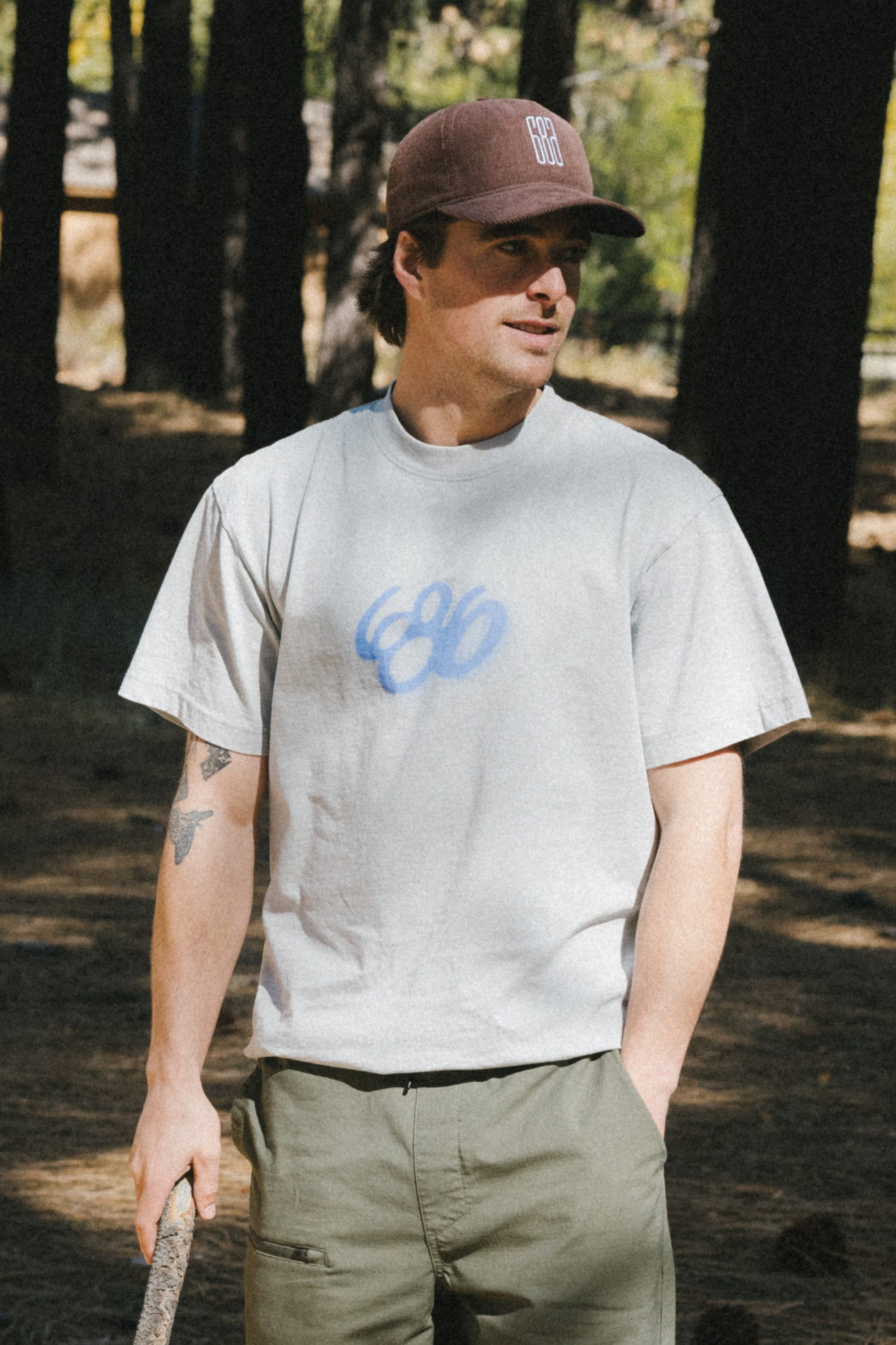 A man in green pants, a brown cap, and the 686 Spray Relaxed SS Tee by 686—a light gray, 100% cotton men's t-shirt with a blue design—stands in a wooded area holding a stick and looking to the side.