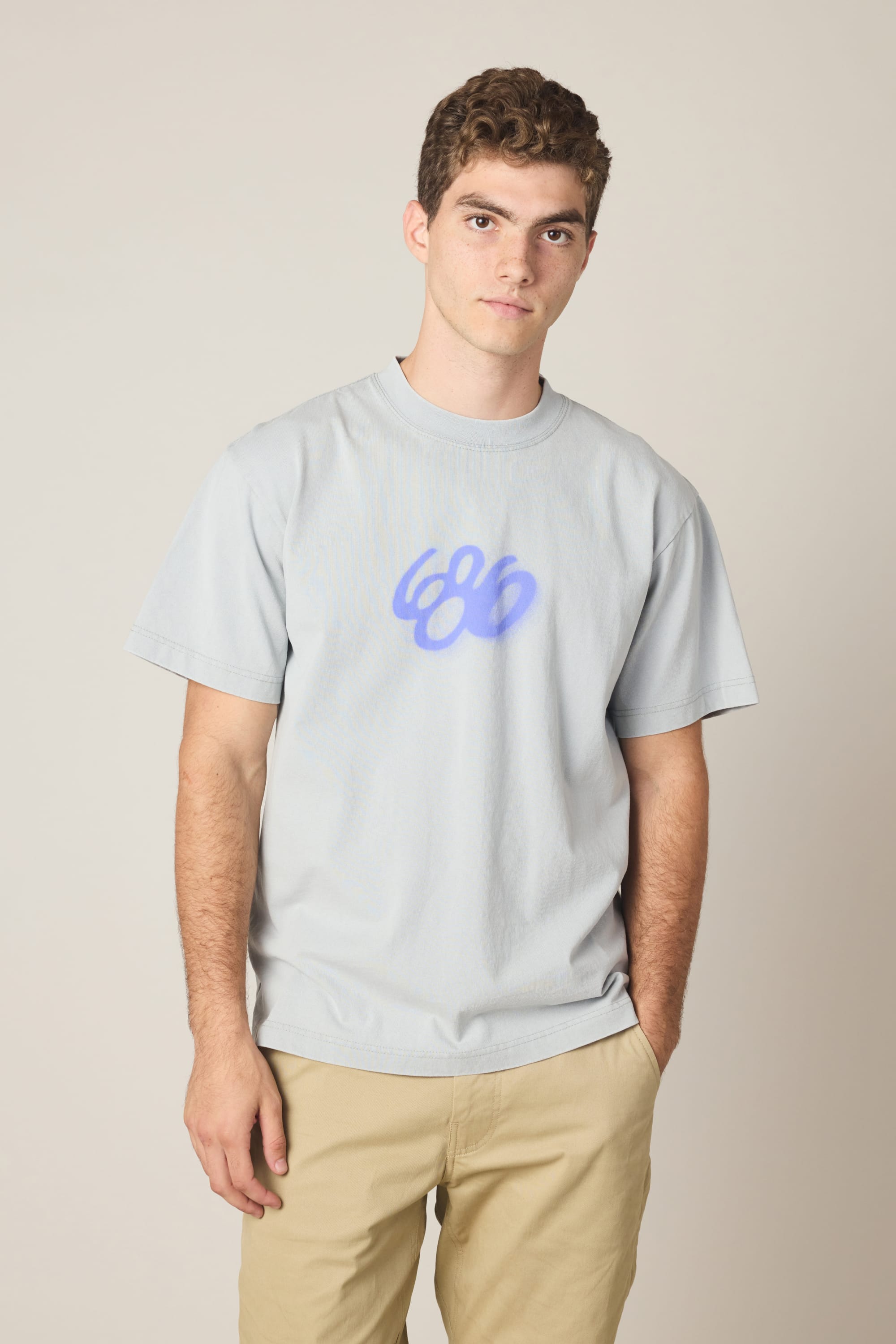 A young man with short curly hair wears the 686 Spray Relaxed SS Tee, a light gray 100% cotton men's t-shirt with a blue abstract 686 logo, and beige pants. He stands facing the camera against a plain light background, expression neutral.