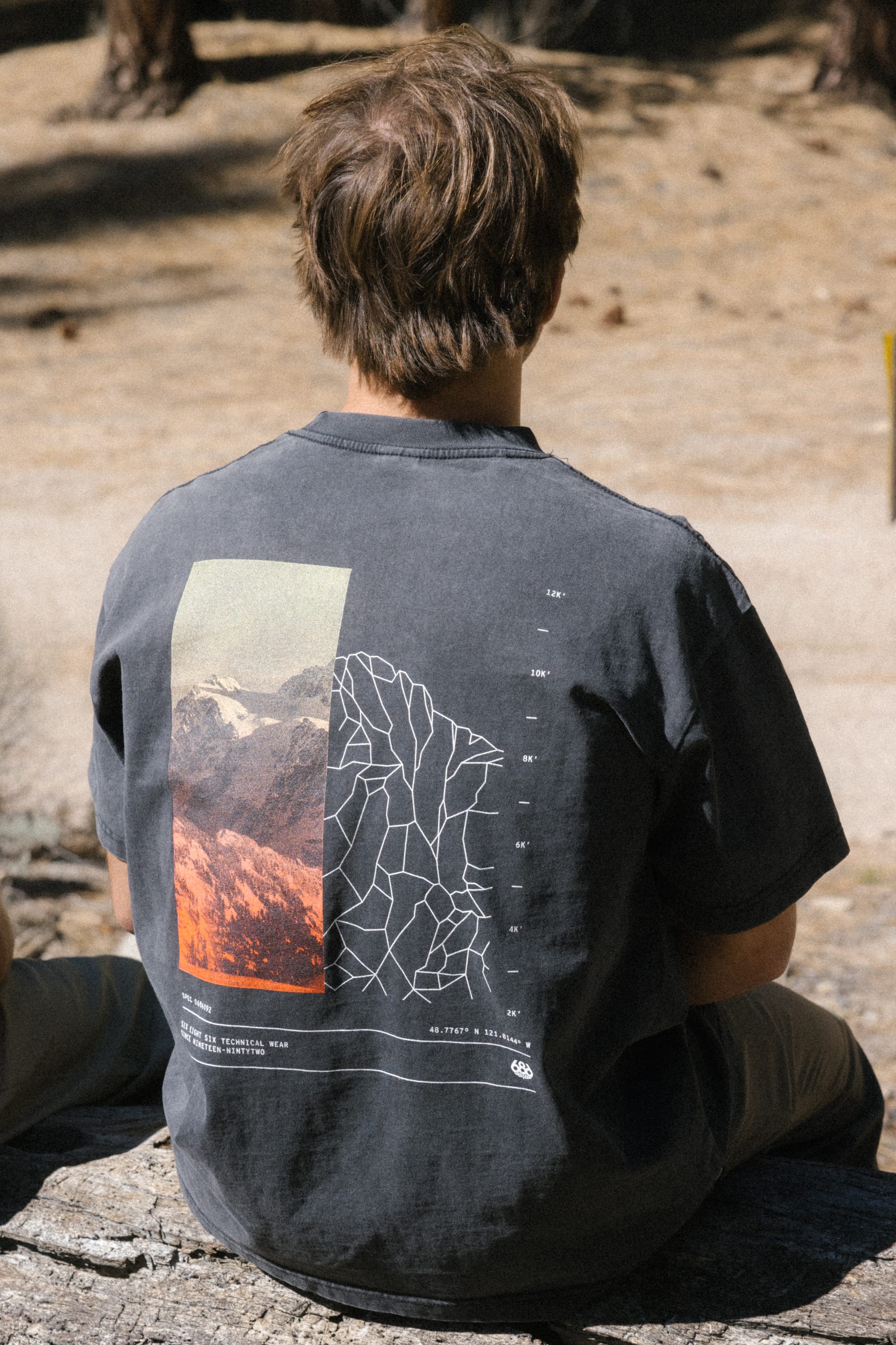 A person with brown hair sits on a rock outdoors, wearing the 686 Peaks Relaxed SS Tee by 686—a men's 100% cotton t-shirt with a mountain and abstract line graphic. The scene is set in a sandy, wooded area.