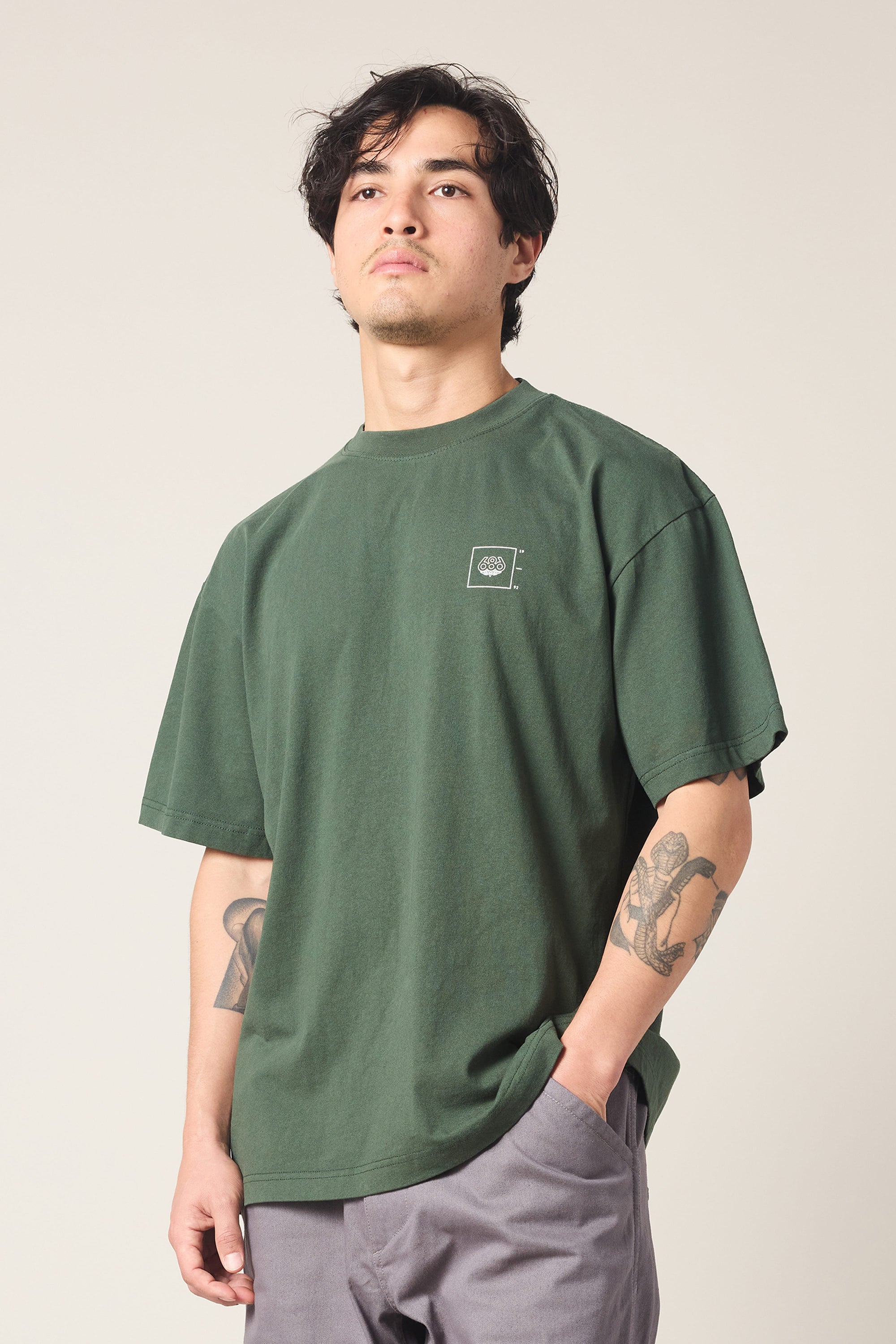 A person with dark hair wears a loose-fitting 686 Peaks Relaxed SS Tee by 686 in forest green and gray pants, standing with one hand in their pocket. Both arms have visible tattoos against a plain, light background.