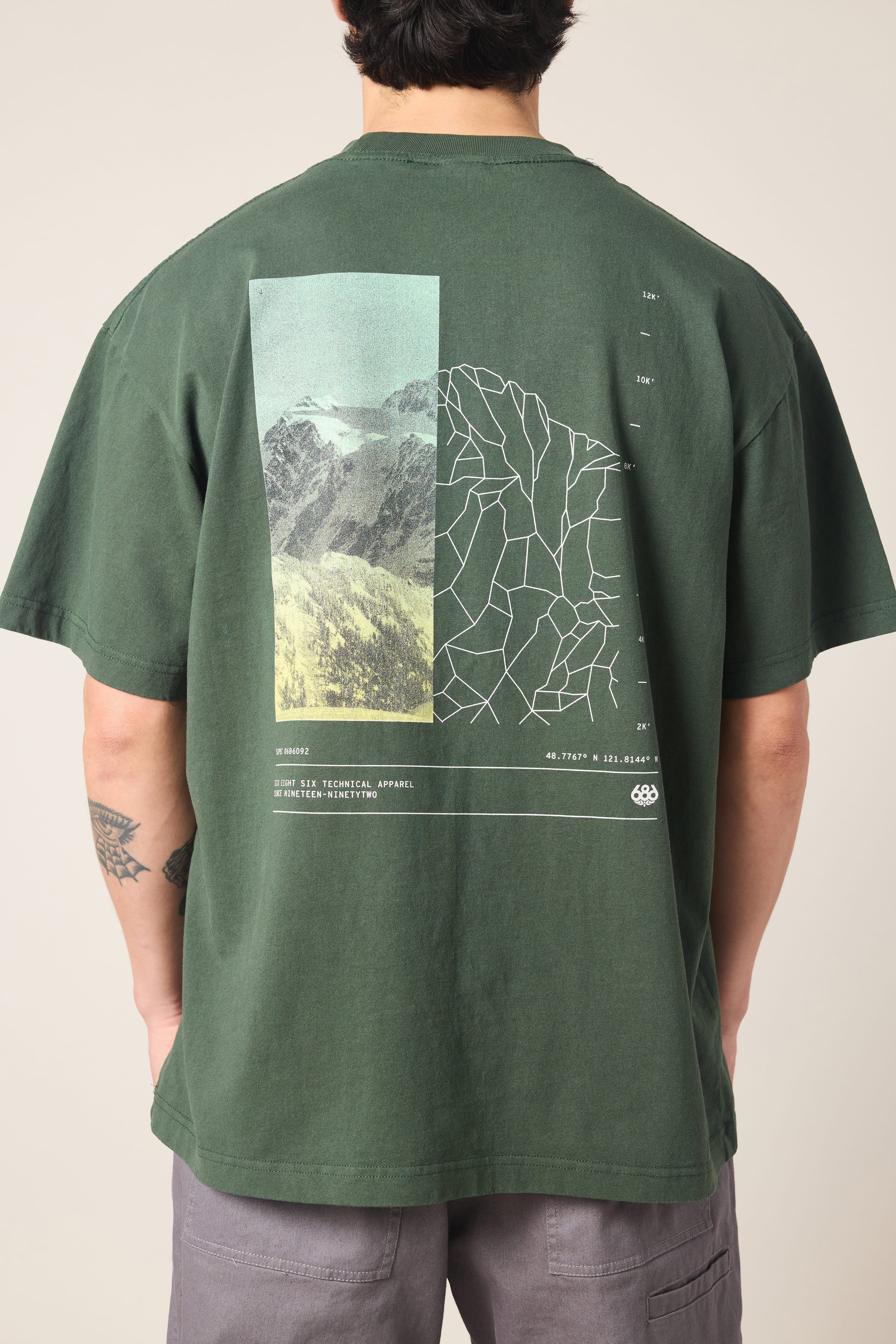 A person stands with their back to the camera, wearing the 686 Peaks Relaxed SS Tee by 686—a men's dark green 100% cotton t-shirt featuring a mountain photo, geometric line design, and small white text graphic on the back.