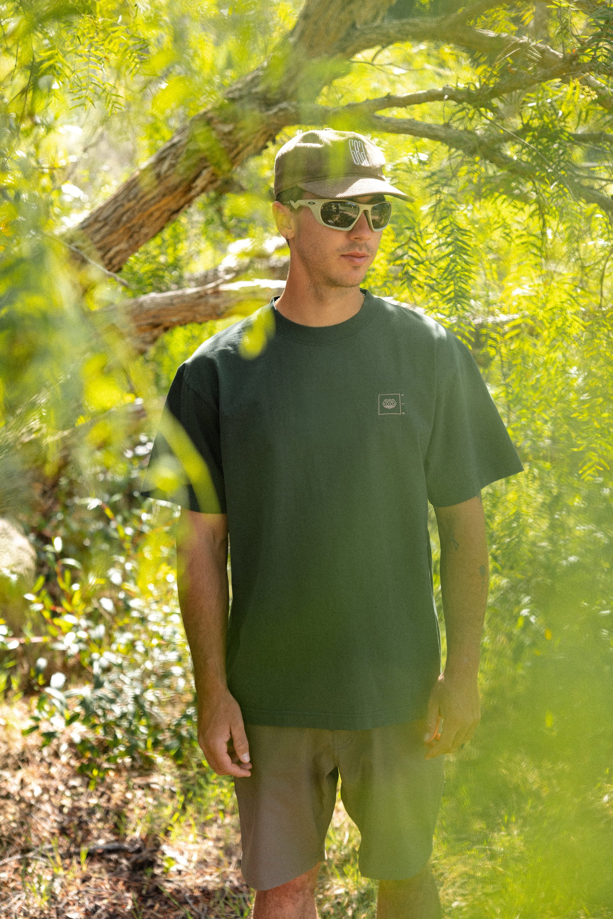A man wearing sunglasses, shorts, a cap, and the 686 Peaks Relaxed SS Tee by 686 stands outdoors amid lush greenery with sunlight filtering through the trees.