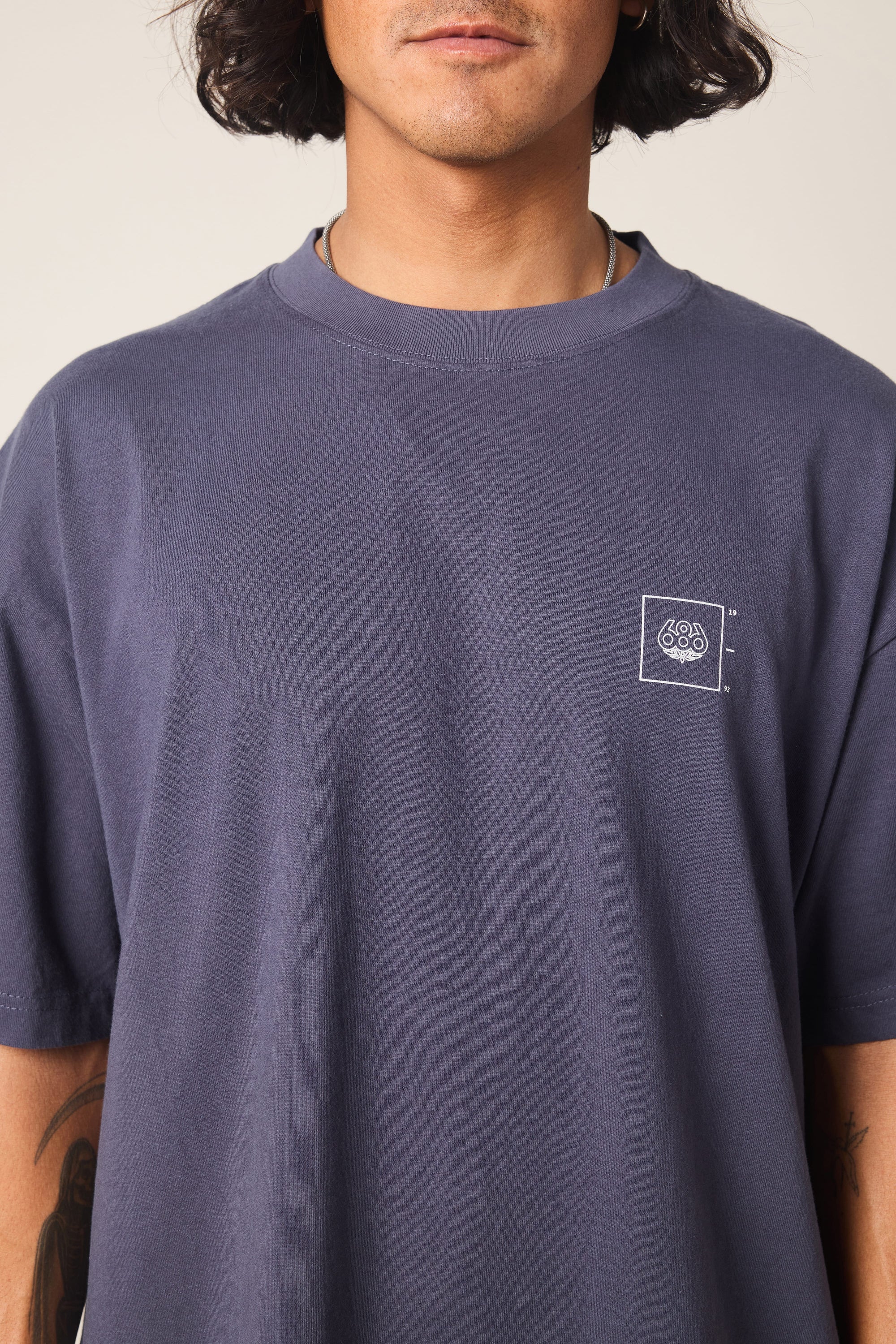 A person with medium-length dark hair wears the 686 Peaks Relaxed SS Tee by 686—a loose-fitting, faded purple men's T-shirt made from 100% cotton with a small white geometric logo on the left chest. Photo cropped nose to waist.