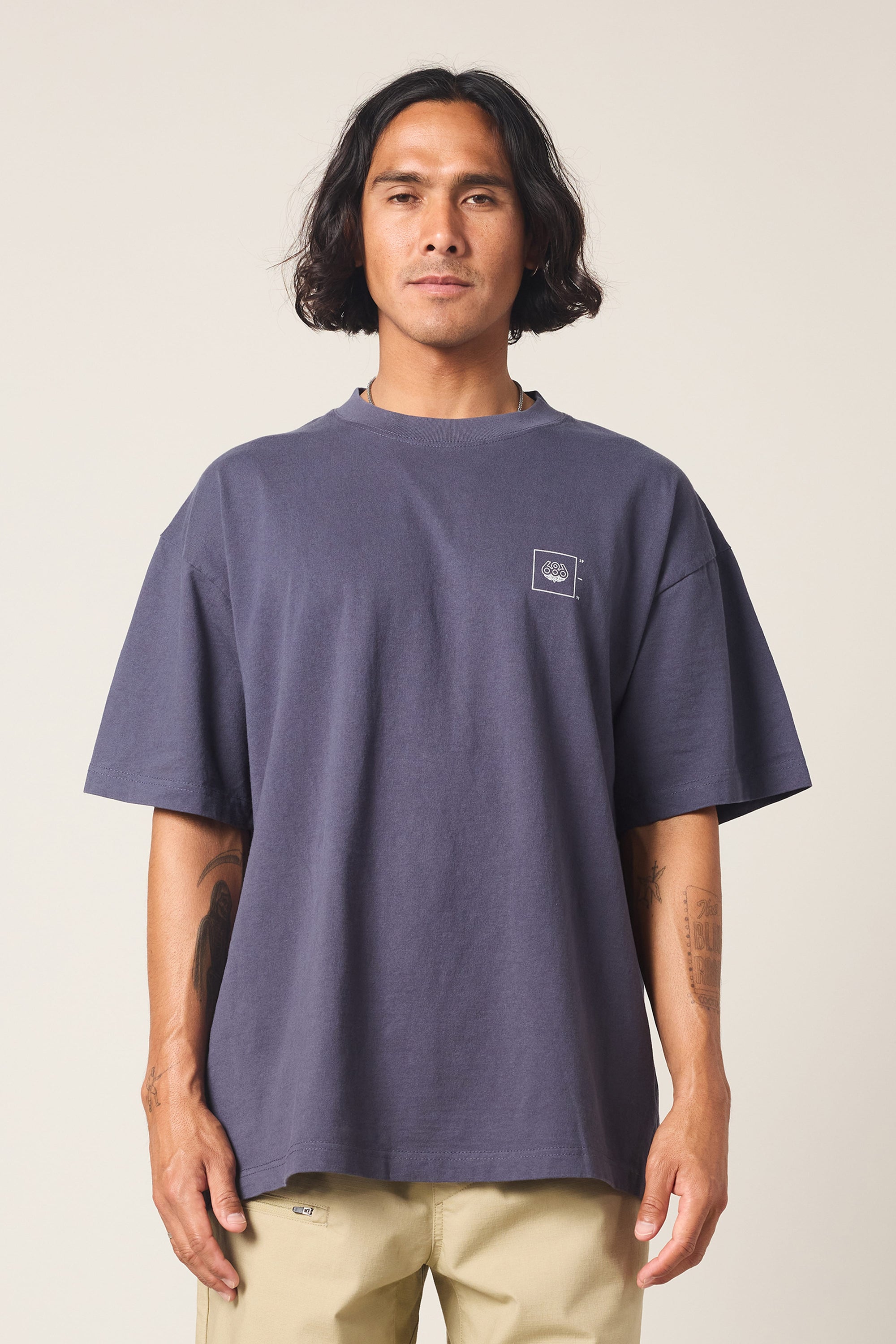 A person with medium-length dark hair wears a loose-fitting 686 Peaks Relaxed SS Tee in dark blue and light beige pants, standing against a plain light background, facing the camera with a neutral expression.