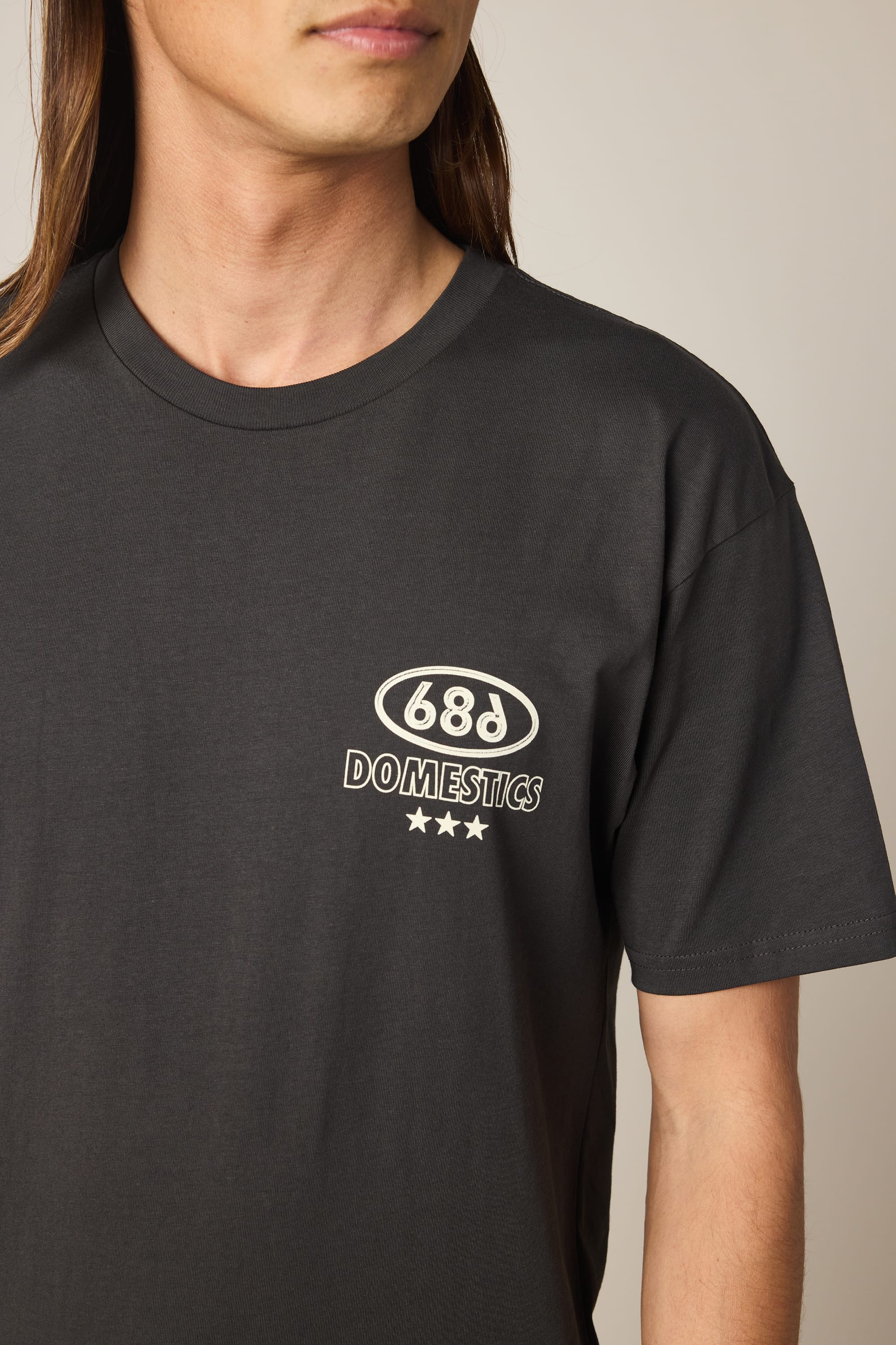 A person with long hair wears the 686 Men's Domestics Authentic SS Tee, a black 100% cotton shirt featuring the 686 logo and three white stars on the upper left chest. The background is plain and neutral.