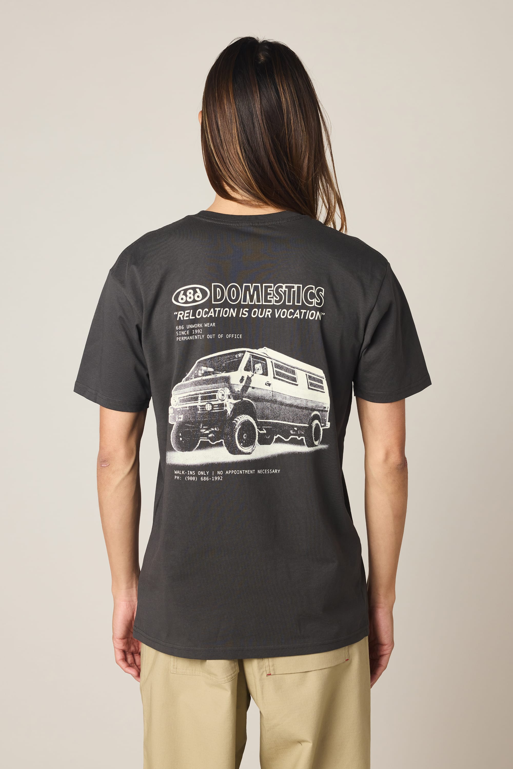 A person with long hair, facing away from the camera, wears a black 686 Men's Domestics Authentic SS Tee featuring a white van graphic and "688 Domestics, Relocation is Our Vocation" text on the back. Brand: 686.