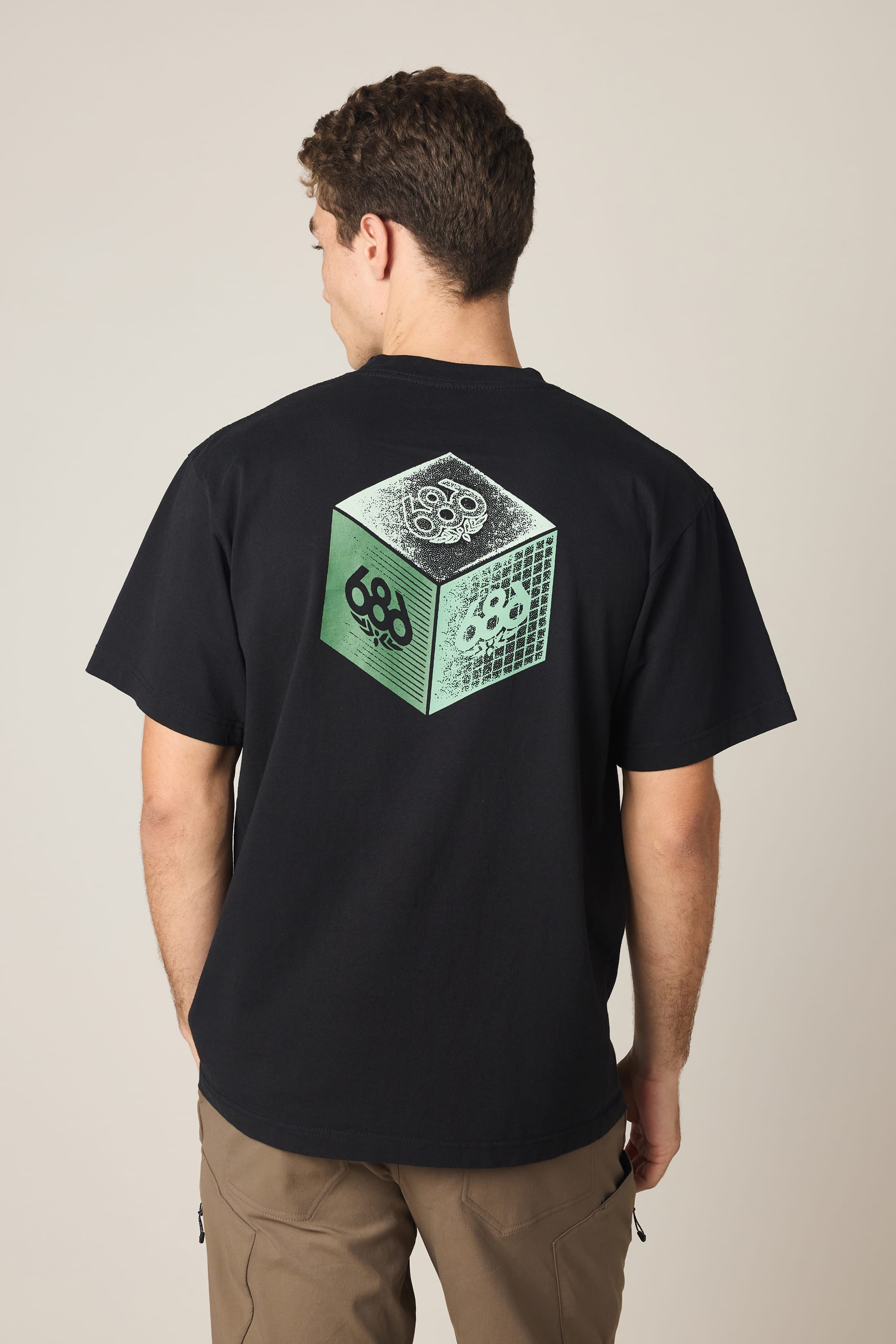 A person with short brown hair, seen from behind, wears the 686 Men's Unit Relaxed SS Tee—a black 100% cotton T-shirt by 686 featuring a green and white cube graphic with skull designs on the back. He is also wearing brown pants.