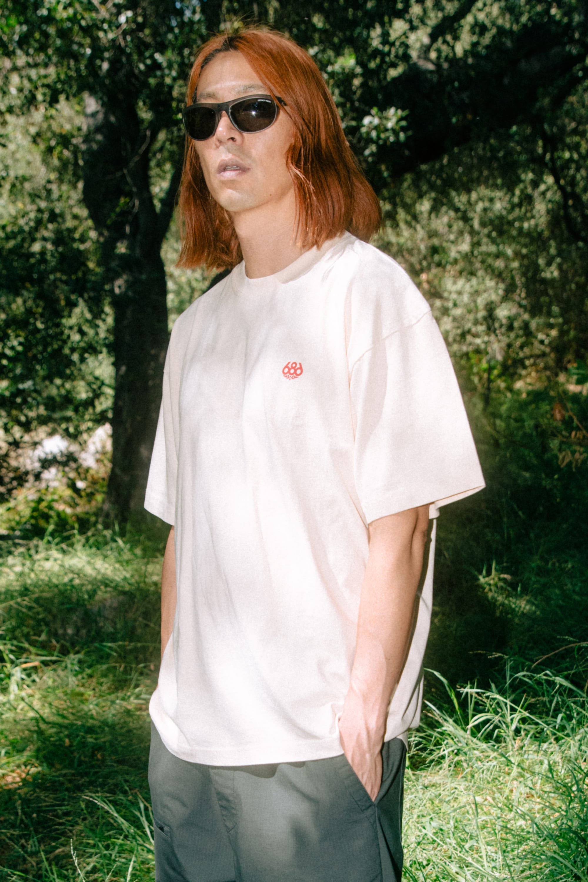 A person with shoulder-length red hair wearing sunglasses and the 686 Men's Unit Relaxed SS Tee by 686, paired with gray shorts, stands outdoors in a sunlit, wooded area with green grass and trees in the background.