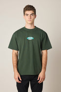 A young man with short curly hair wears the 686 Men's Jib Relaxed SS Tee by 686, a dark green 100% cotton t-shirt with a small colorful oval chest logo, paired with black pants, standing against a plain light background.