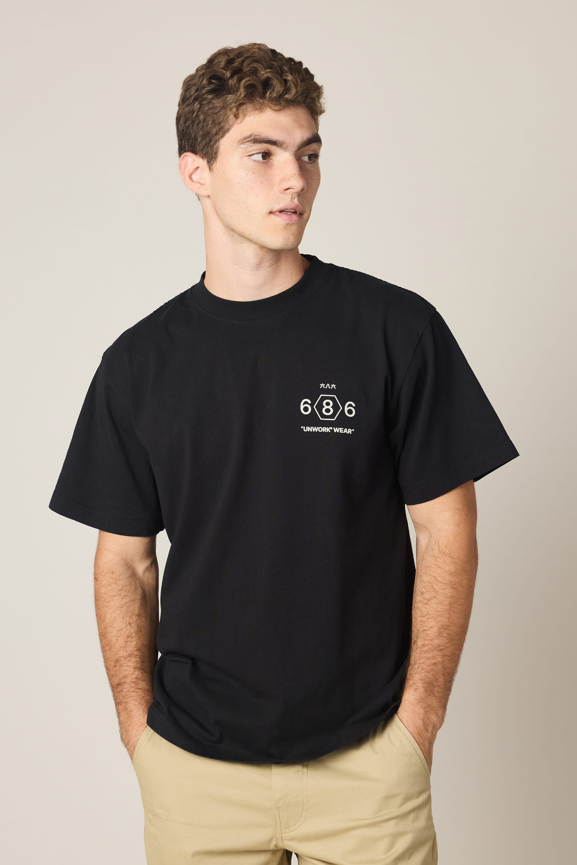 A young man with short curly hair wears the 686 Men's Unwork Spec Relaxed SS Tee, a black 100% cotton t-shirt featuring a small white 686 logo and text on the left chest. He stands with hands in his beige pants pockets, looking to the side.