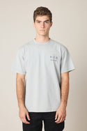 A young man with short curly hair wears the 686 Men's Unwork Spec Relaxed SS Tee in light gray and black pants, standing against a plain background. The 100% cotton tee by 686 features small printed text and numbers on the left chest.
