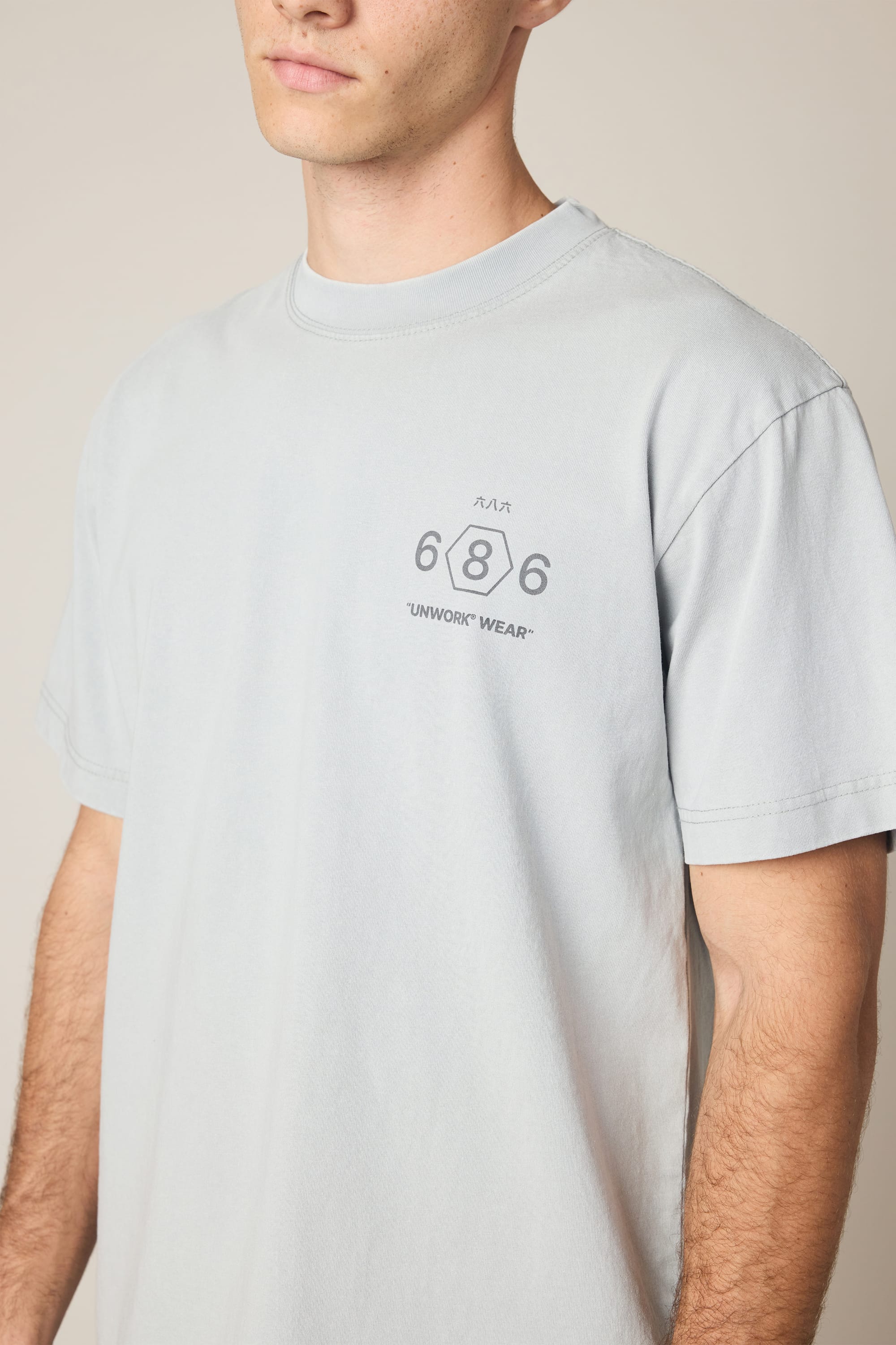 A person is wearing the 686 Men's Unwork Spec Relaxed SS Tee by 686, a light gray 100% cotton t-shirt featuring "686 UNWORK WEAR" and a small graphic printed on the left chest. The person's face is partially out of frame.