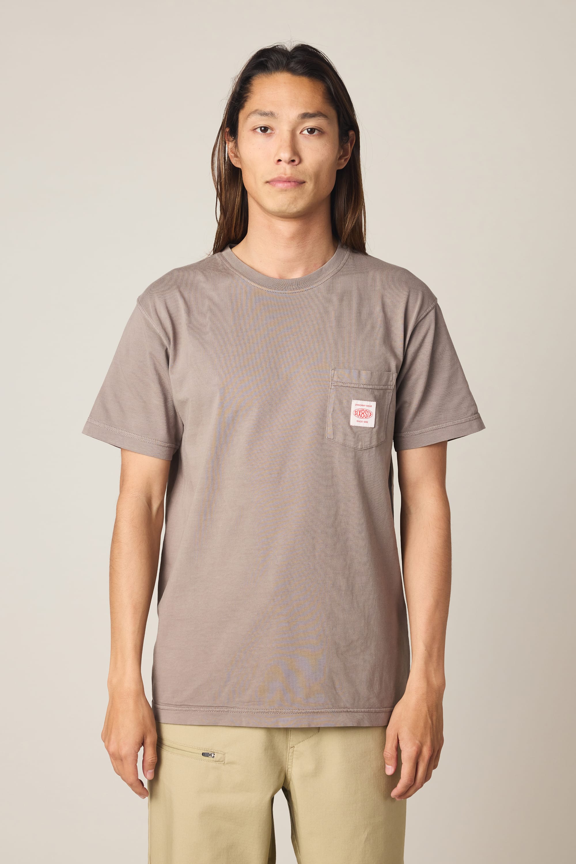 A person with long hair stands facing forward, wearing the 686 Men's Unwork Authentic SS Pocket Tee by 686—a taupe 100% cotton t-shirt featuring a small patch on the chest pocket—paired with light beige pants. The background is plain and neutral.