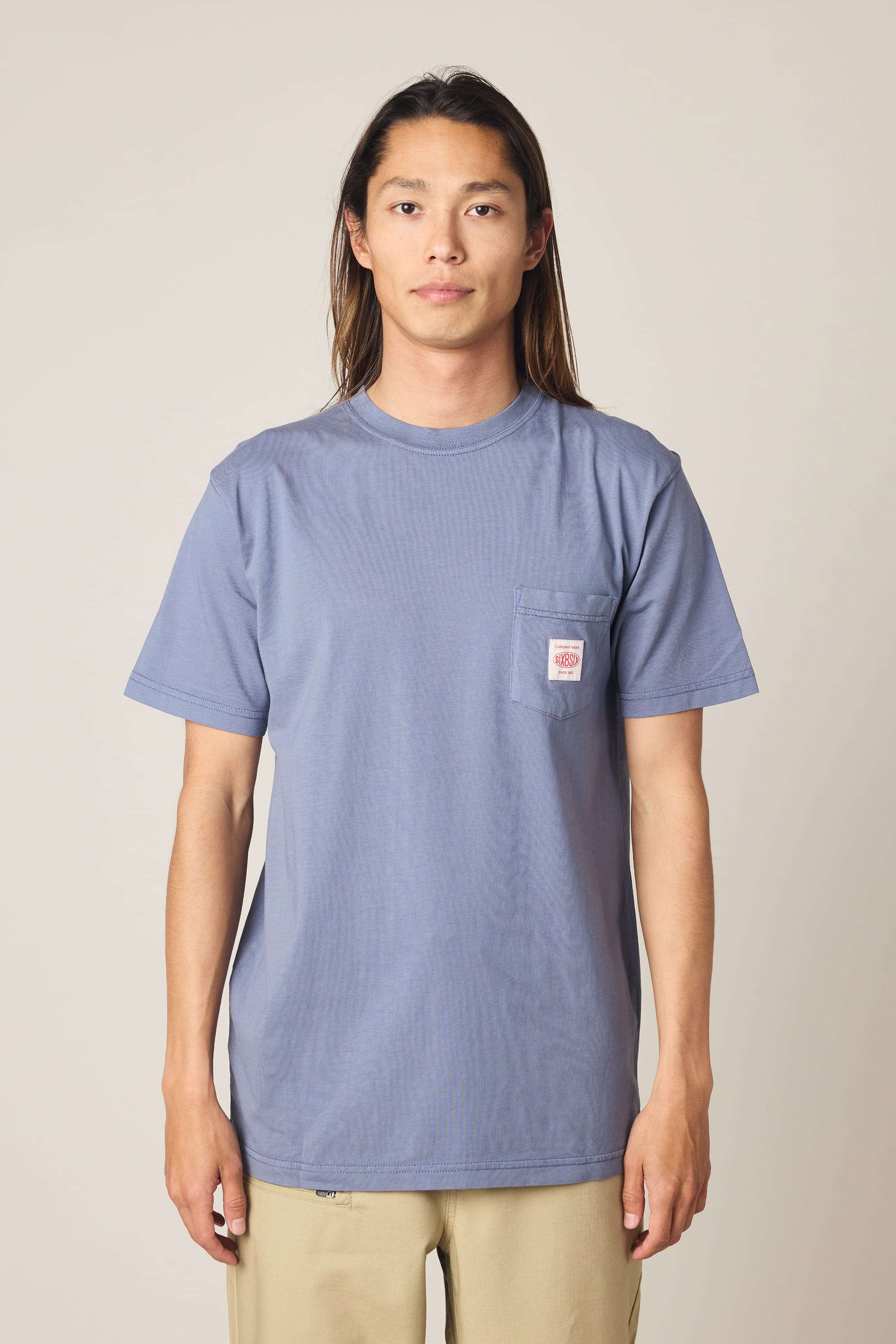 A person with long hair wears the 686 Men's Unwork Authentic SS Pocket Tee in blue—made from 100% cotton with a chest pocket—paired with beige pants, standing against a plain light background.