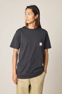 A person with long hair wears the 686 Men's Unwork Authentic SS Pocket Tee in dark grey and beige pants, standing slightly turned against a plain background.