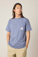 A person with long hair wears the 686 Men's Unwork Authentic SS Pocket Tee, a light blue 100% cotton shirt with a chest pocket, along with beige pants, standing hands in pockets and looking to the side against a plain background.