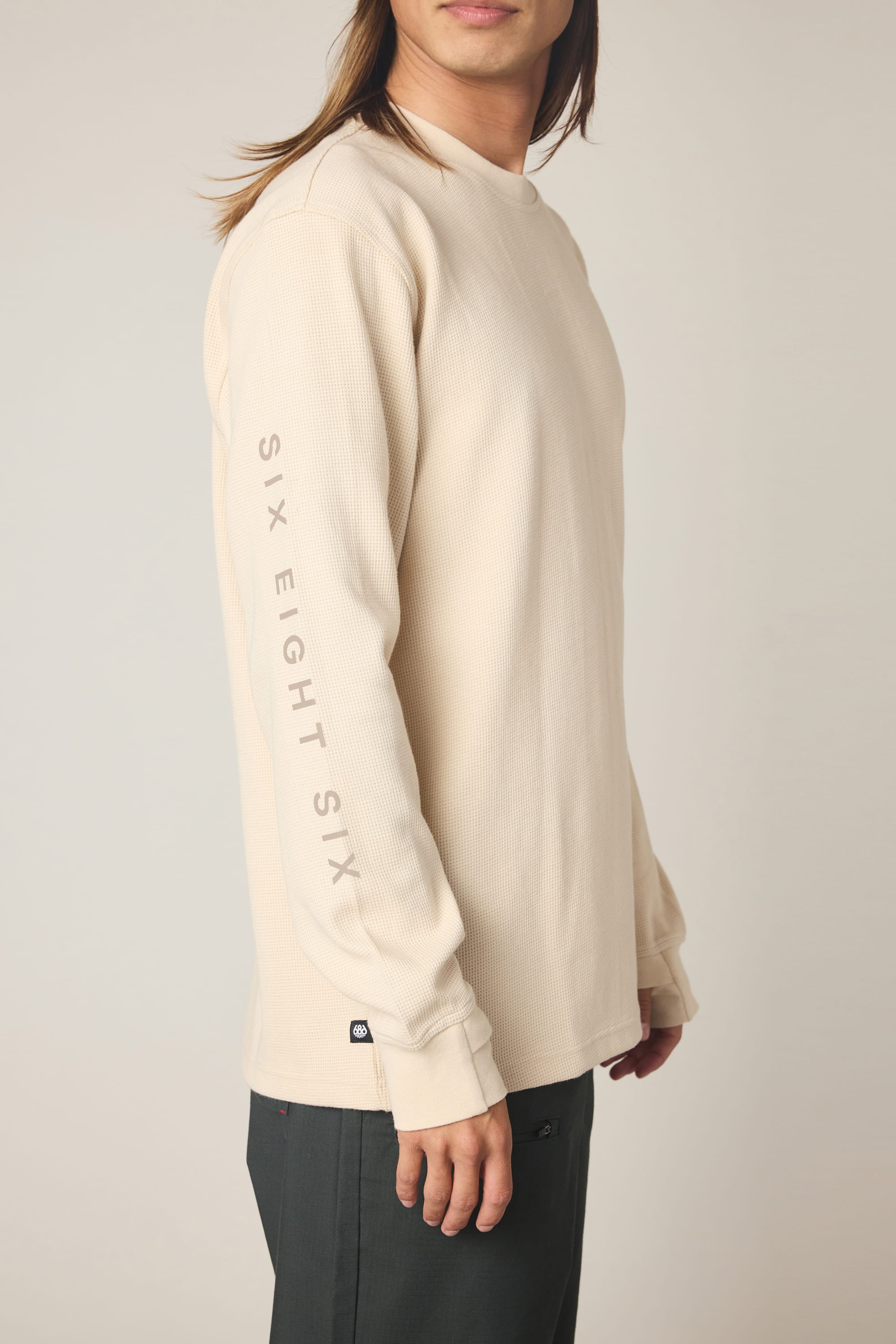 A man wearing the 686 Men's Let's Chill Waffle LS Crew in cream, featuring "686" printed vertically on the left sleeve, stands sideways against a plain light background.