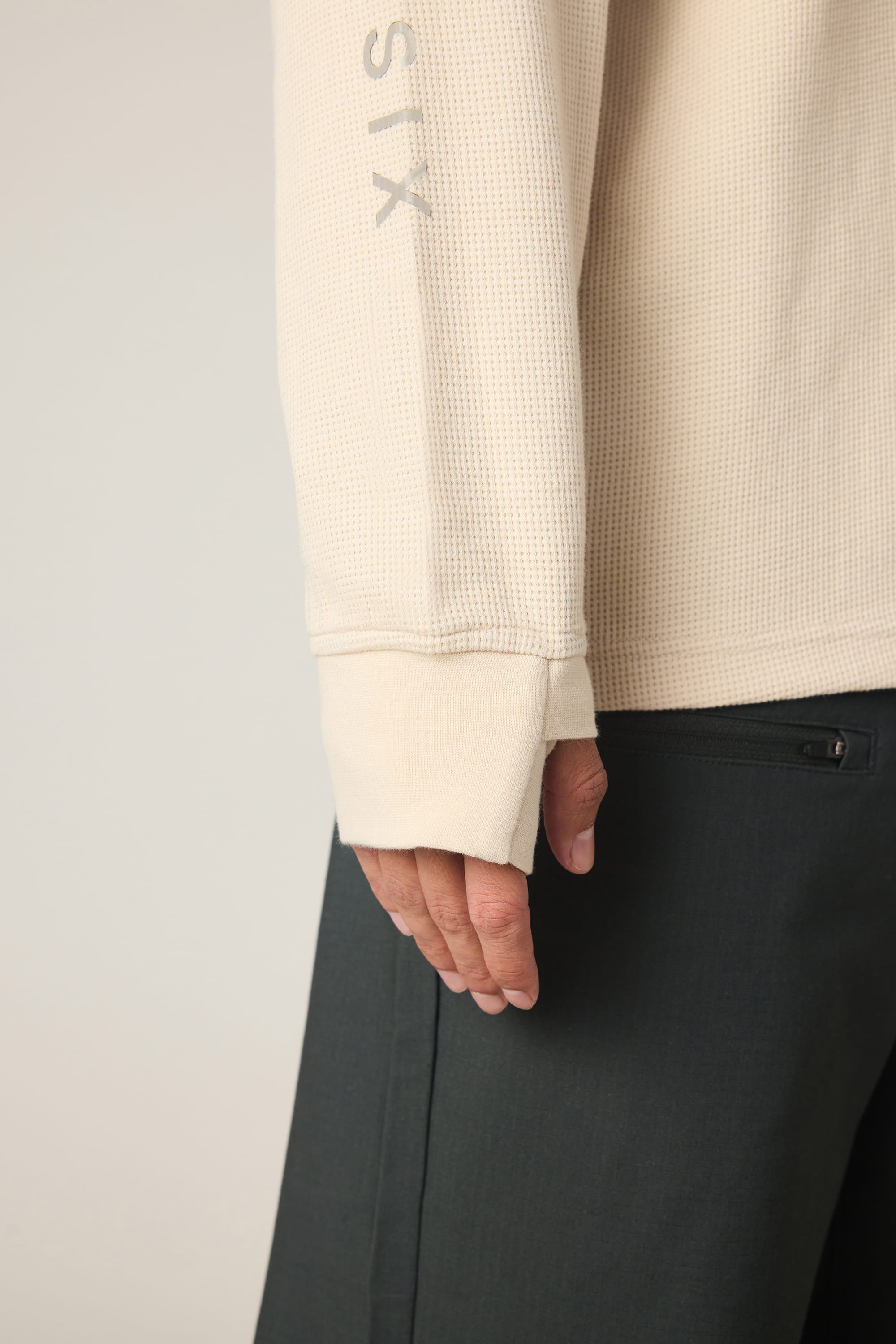 A close-up of a person’s arm and hand wearing the 686 Men's Let's Chill Waffle LS Crew in cream with thumbhole detail, paired with dark pants featuring a visible back pocket and zipper.