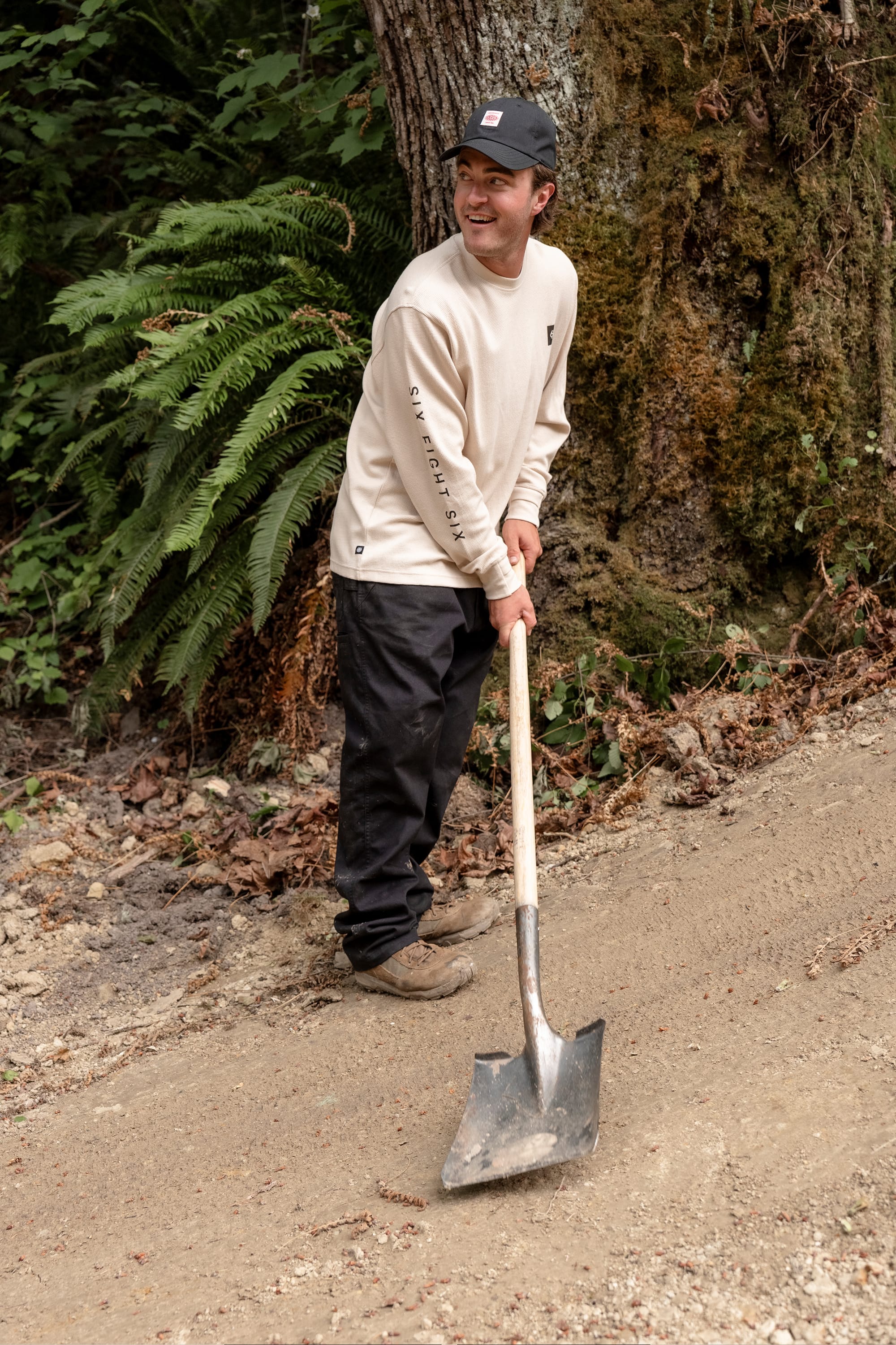A person wearing the 686 Men's Let's Chill Waffle LS Crew in beige, along with black pants and a black cap, is smiling while holding a shovel and standing on a dirt path surrounded by ferns and trees in a forested area.