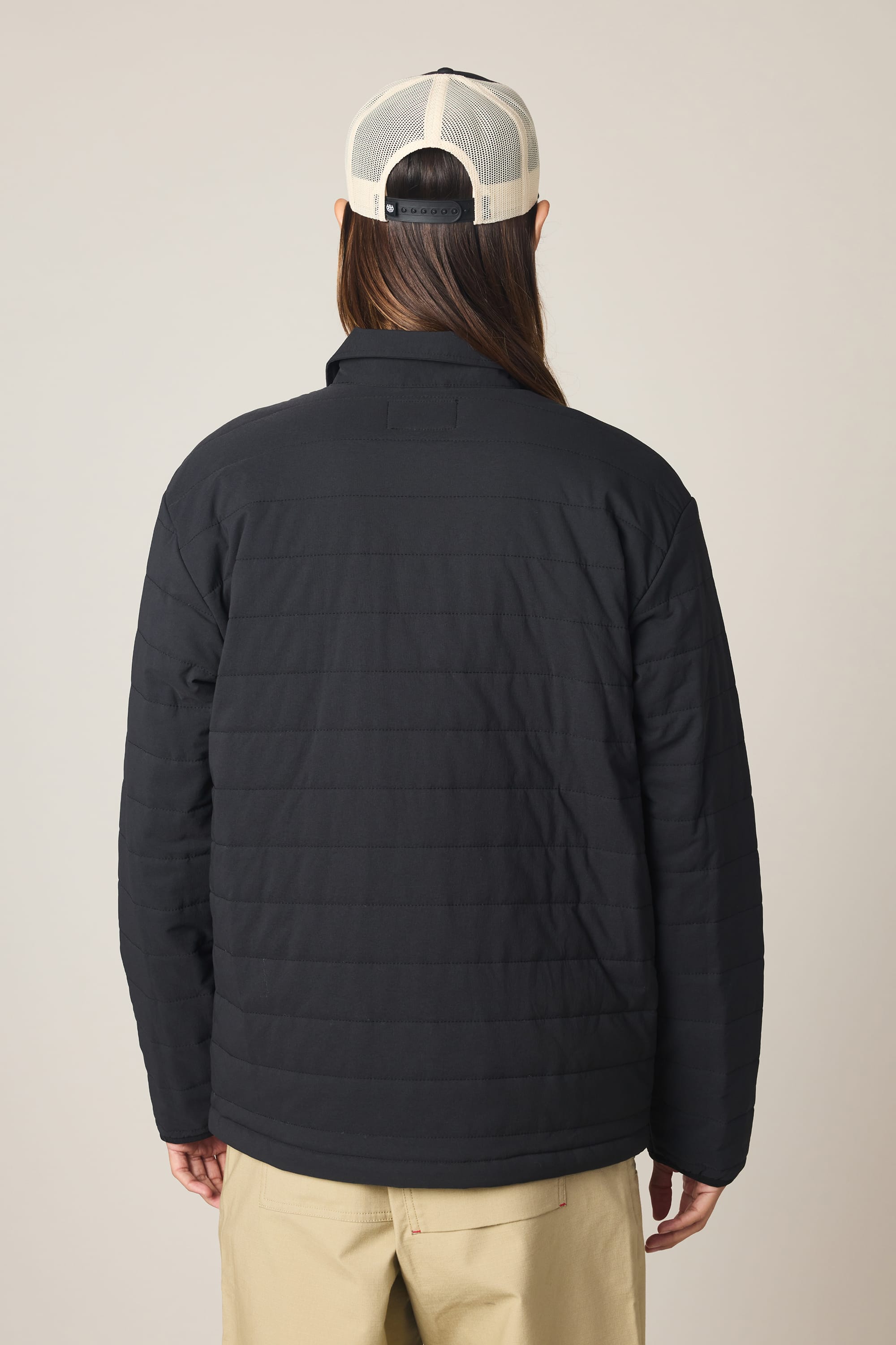 A person with long hair, seen from behind against a plain light background, is wearing a white mesh-back cap, beige pants, and the 686 Men's Thermaldry Insulated Shacket by 686.