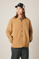 A person with long hair wears the 686 Men's Thermaldry Insulated Shacket by 686 in tan, dark pants, and a black and white baseball cap, standing with hands in jacket pockets and looking to the side against a plain background.
