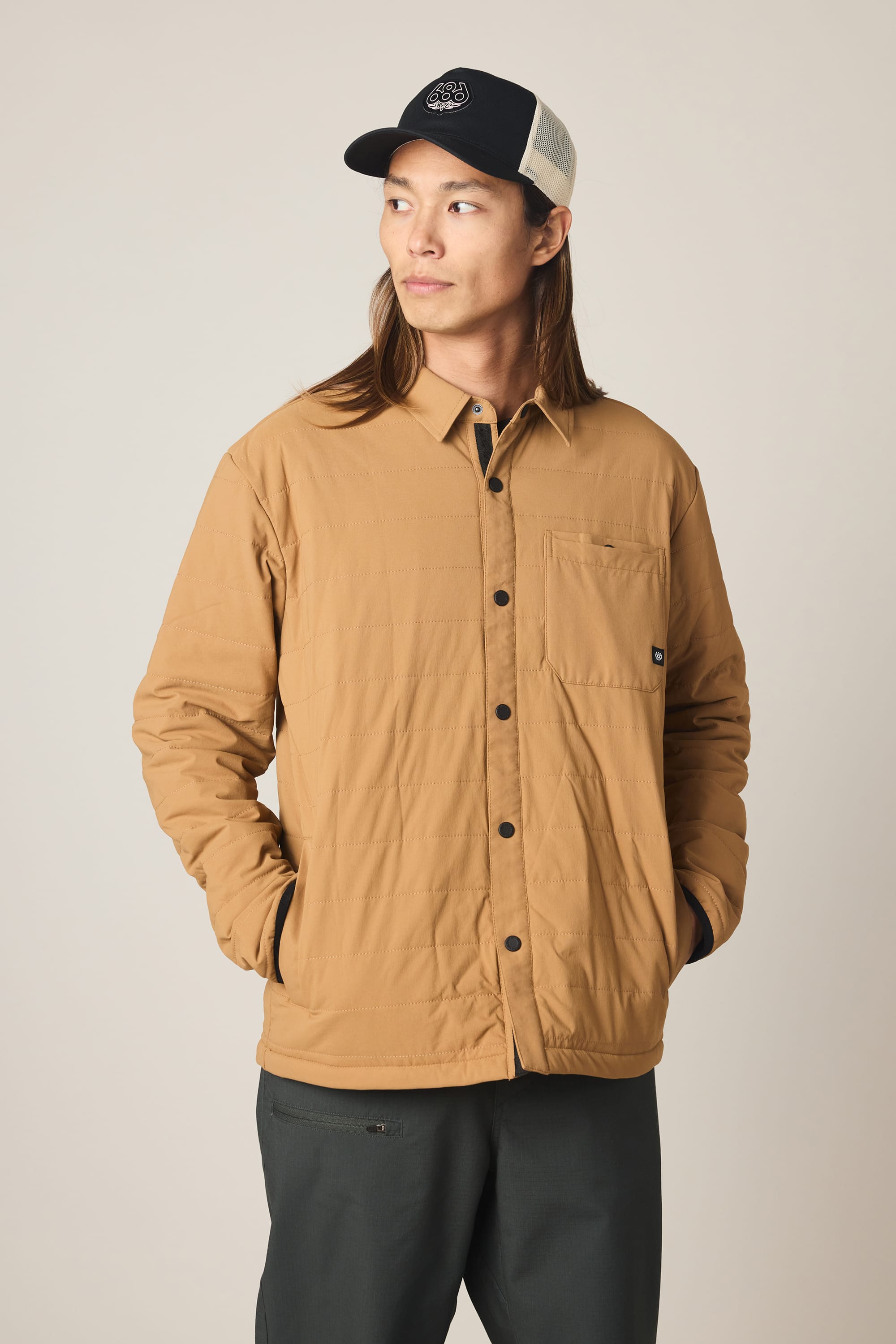 A person with long hair wears the 686 Men's Thermaldry Insulated Shacket by 686 in tan, dark pants, and a black and white baseball cap, standing with hands in jacket pockets and looking to the side against a plain background.