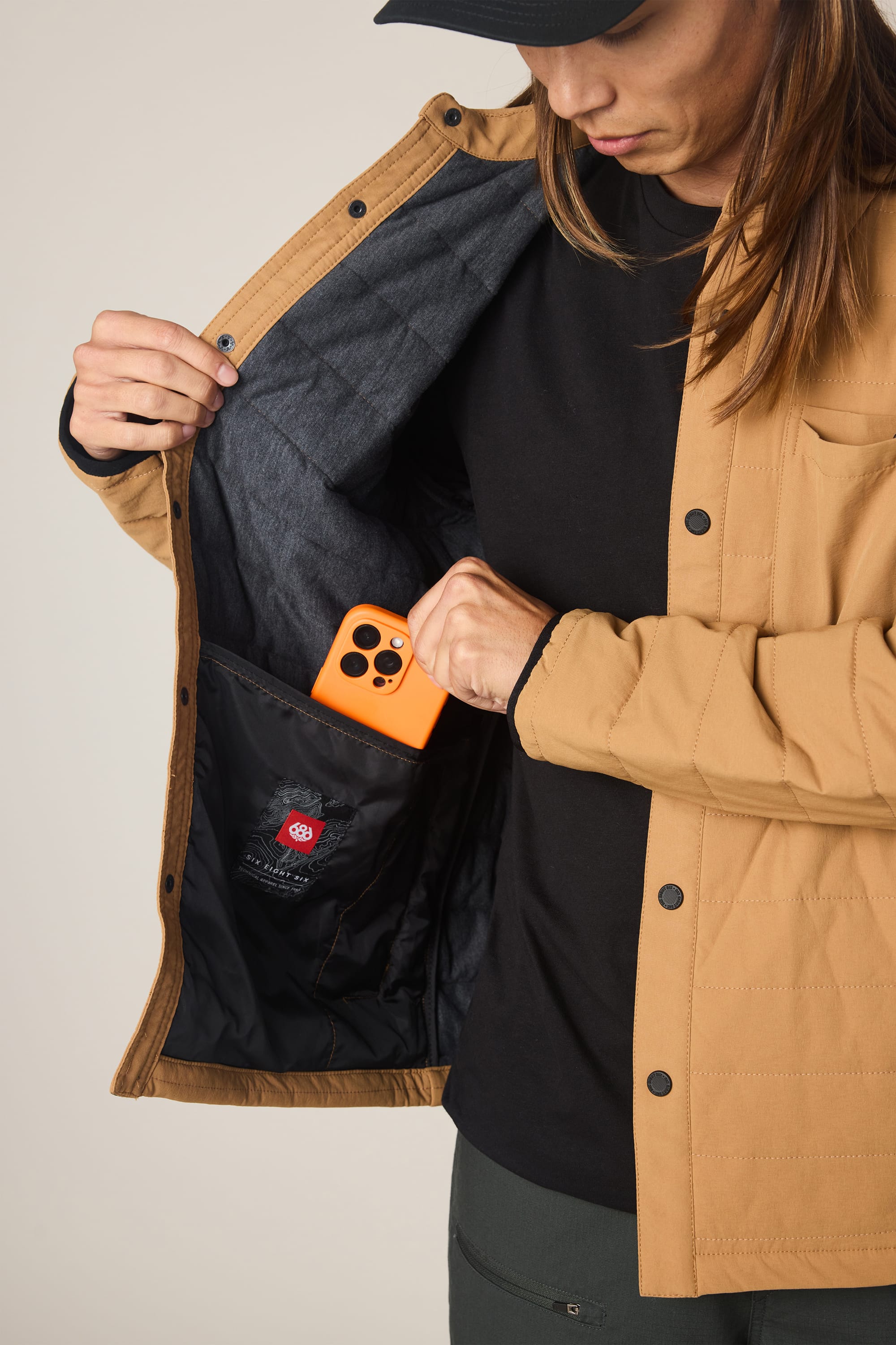 A person wearing the 686 Men's Thermaldry™ Insulated Shacket in tan with a black shirt places an orange phone into an inner pocket, showing off the lining and the interior black 686 label.