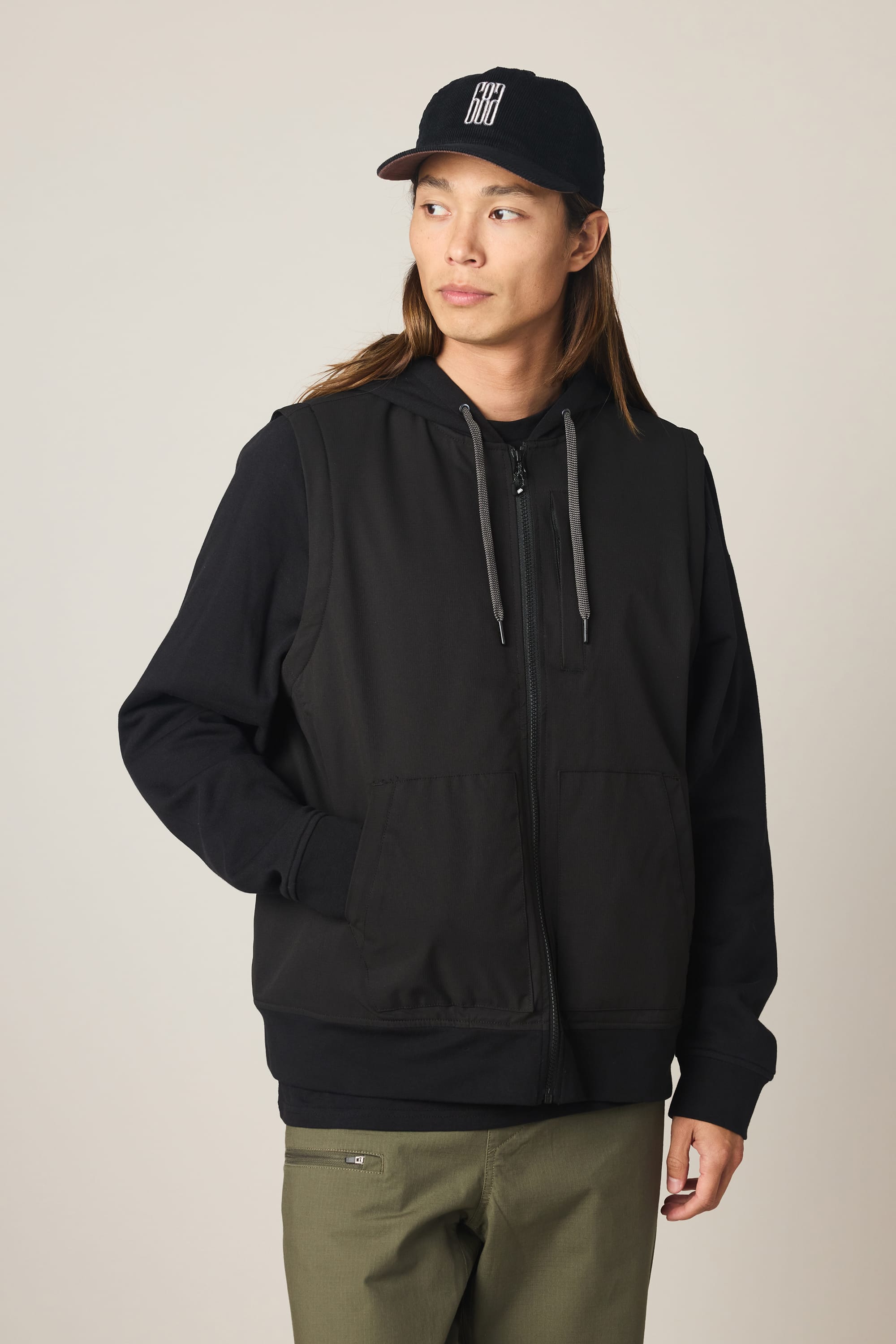 A person with long hair wears the 686 Men's Merge Quilted Hoody—a black zip-up hoodie with gray drawstrings—paired with a black cap with white lettering and olive green pants, standing against a plain light background.