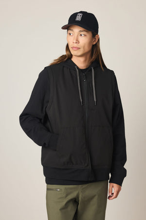 A person wearing a black cap, olive-green pants, and the 686 Men's Merge Quilted Hoody by 686—a black polyester jacket with gray drawstrings—stands against a plain light background, looking slightly to the side with hands in the jacket pockets.