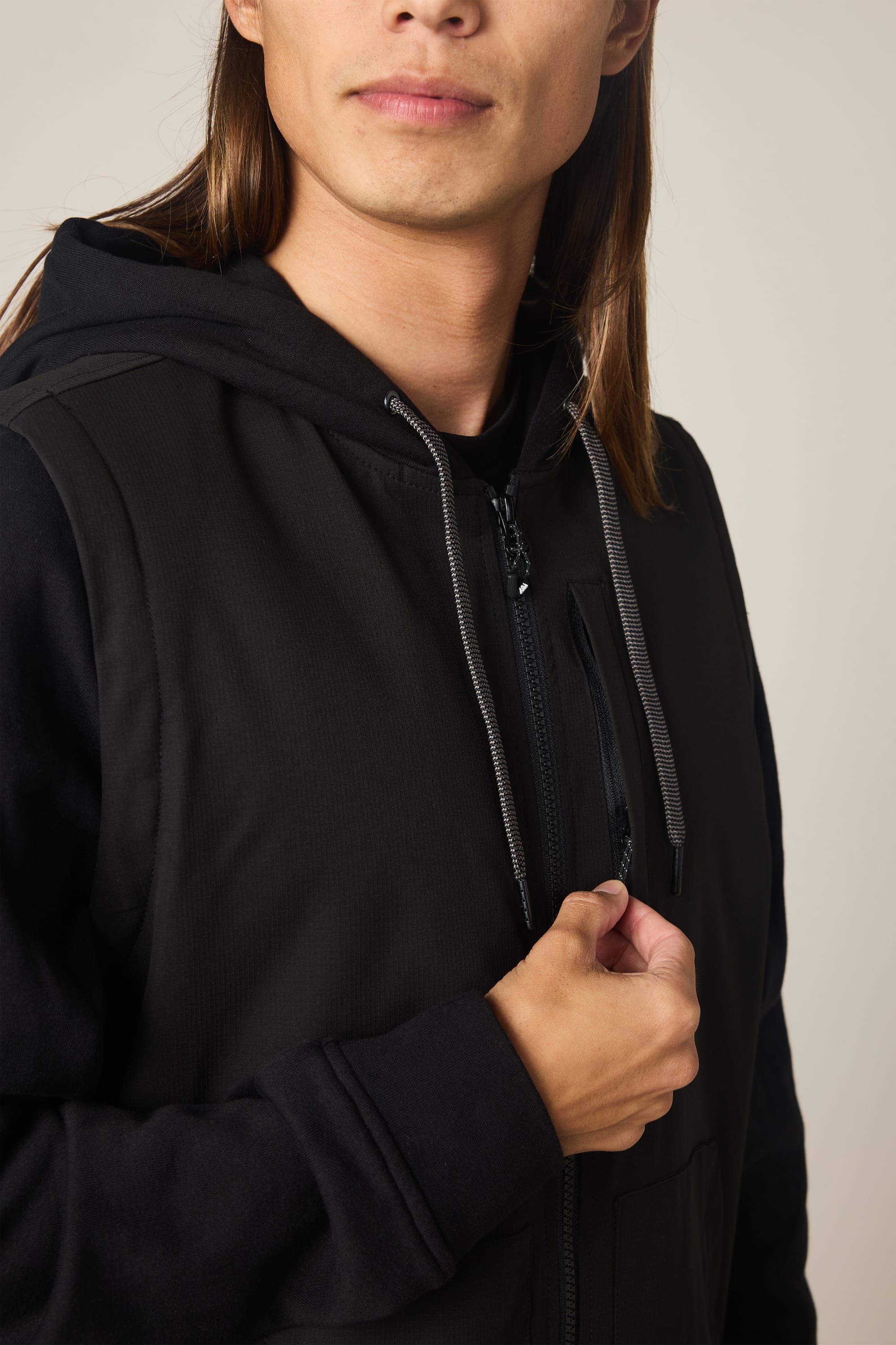 A person with long brown hair wears a 686 Men's Merge Quilted Hoody in black, unzipping the chest pocket and looking slightly to the side. The upper body is shown against a plain background.