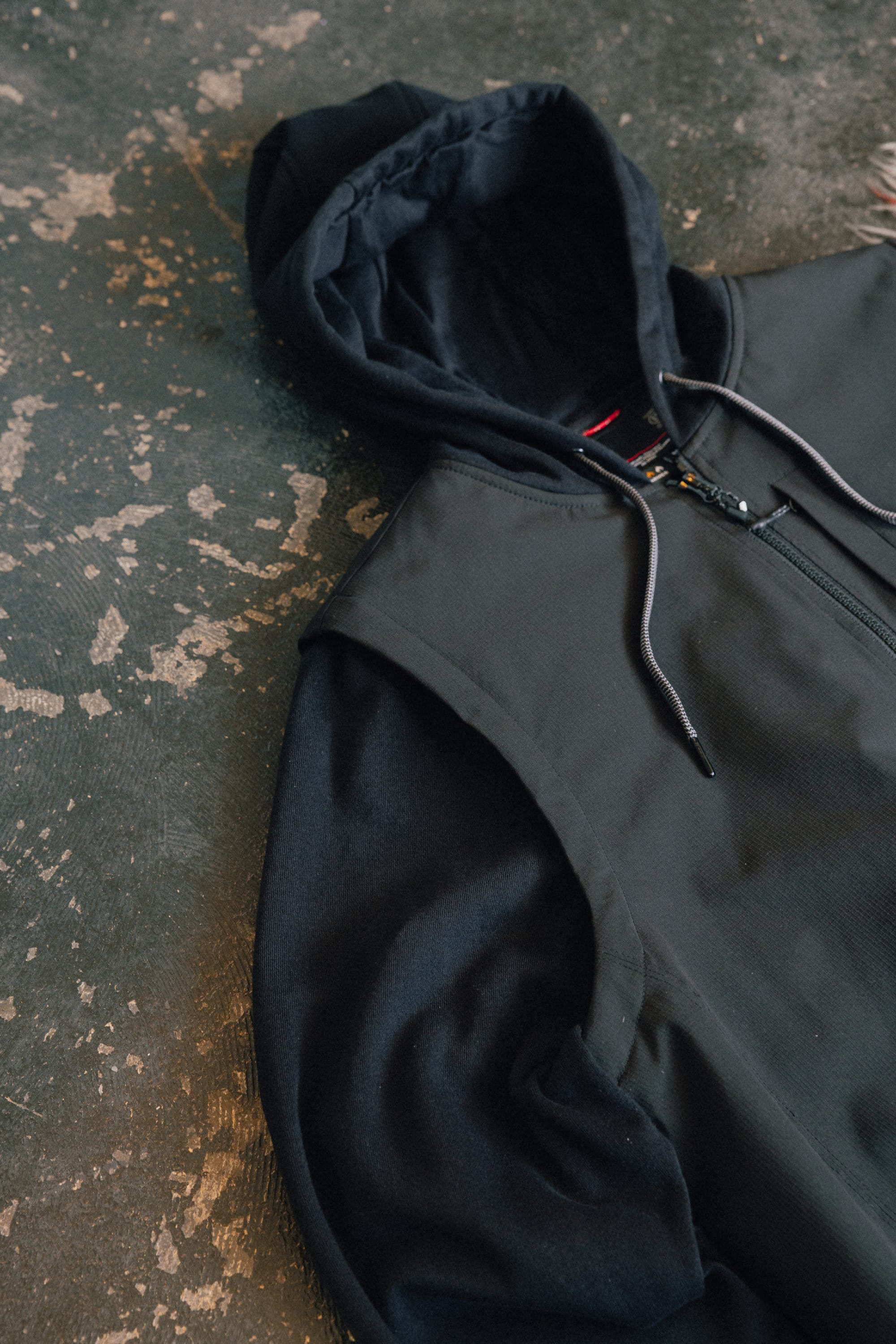 A 686 Men's Merge Quilted Hoody in black, featuring polyester knit construction and a zipper, is laid flat on a textured, worn concrete surface. Only the upper half of the hoody and its hood are visible.