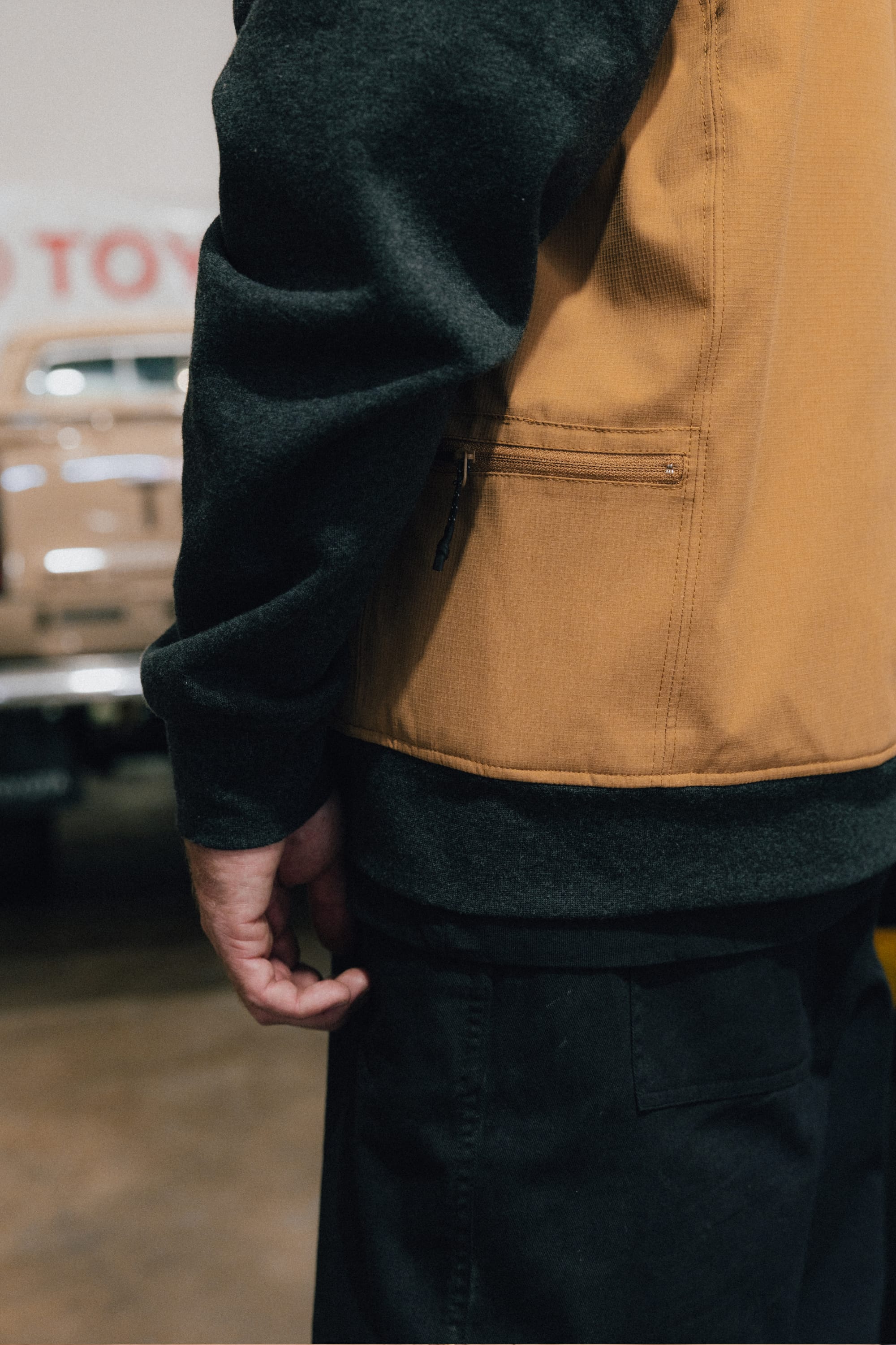A person stands indoors near a tan vintage vehicle, wearing the 686 Men's Merge Quilted Hoody layered under a brown vest. The focus is on their arm and torso, with the background blurred.